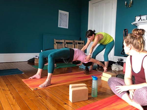 Three women practicing yoga in a cozy, well-lit room with teal walls. One woman is in a plank position on an orange yoga mat, another woman is assisting her, and a third woman is sitting cross-legged on a pink yoga mat, observing.