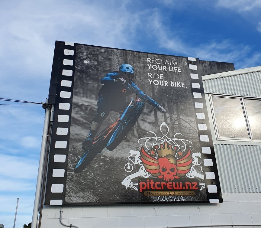 Large outdoor billboard featuring a mountain biker in mid-air with the text "RECLAIM YOUR LIFE. RIDE YOUR BIKE." and a logo with a red skull, wings, and a crown for Pitcrew.nz, a bicycle servicing shop.