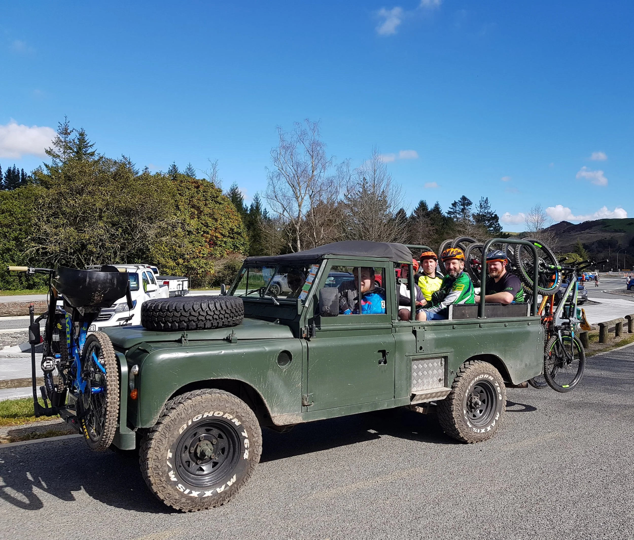 A group of four cyclists with helmets riding in the back of a green vintage Land Rover with bicycles attached to the rear, parked on a paved road near a grassy area with trees and hills under a clear blue sky.