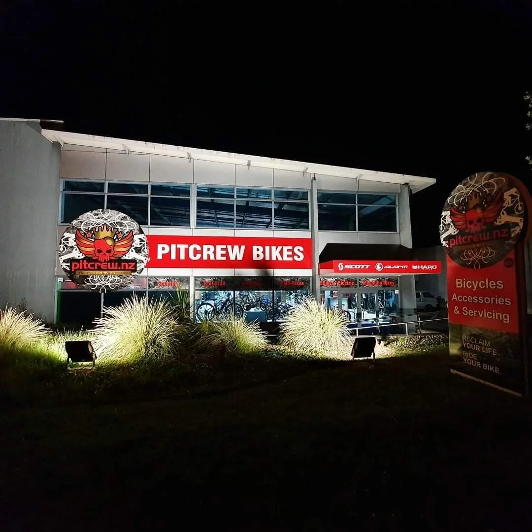 Nighttime view of a bicycle shop called PITCREW BIKES, with large illuminated sign and logo featuring a red skull with wings, situated in a modern building with large glass windows. There is a sign describing bicycles, accessories, and servicing, and