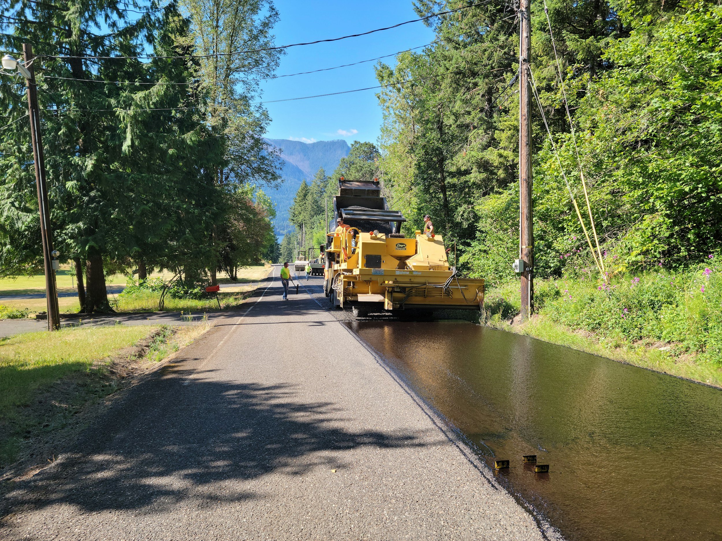 Road construction workers paving a new road with asphalt, surrounded by trees and power lines, with mountains in the background on a clear, sunny day.
