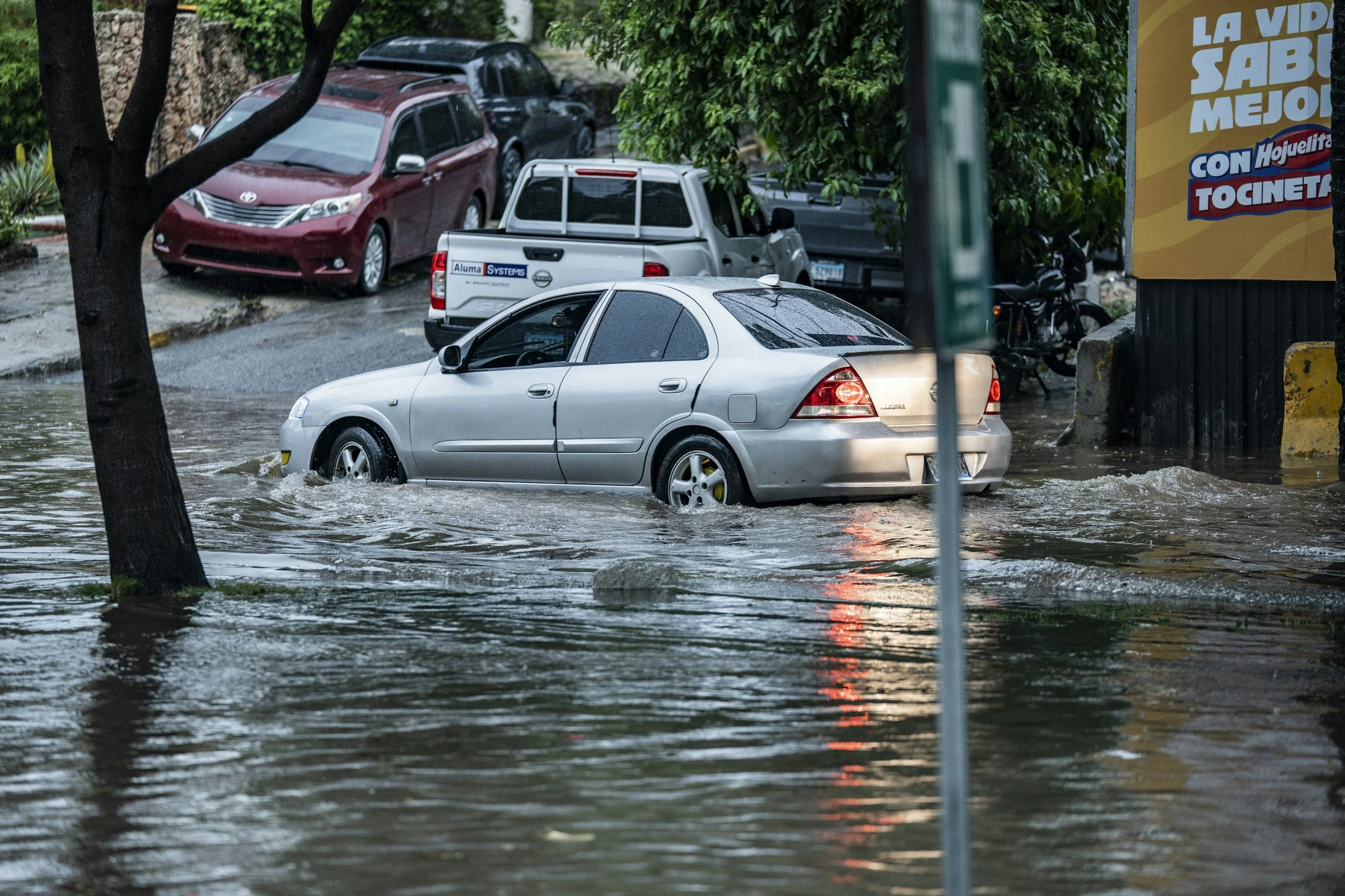 A silver sedan partially submerged in floodwater with trees and parked cars in the background.