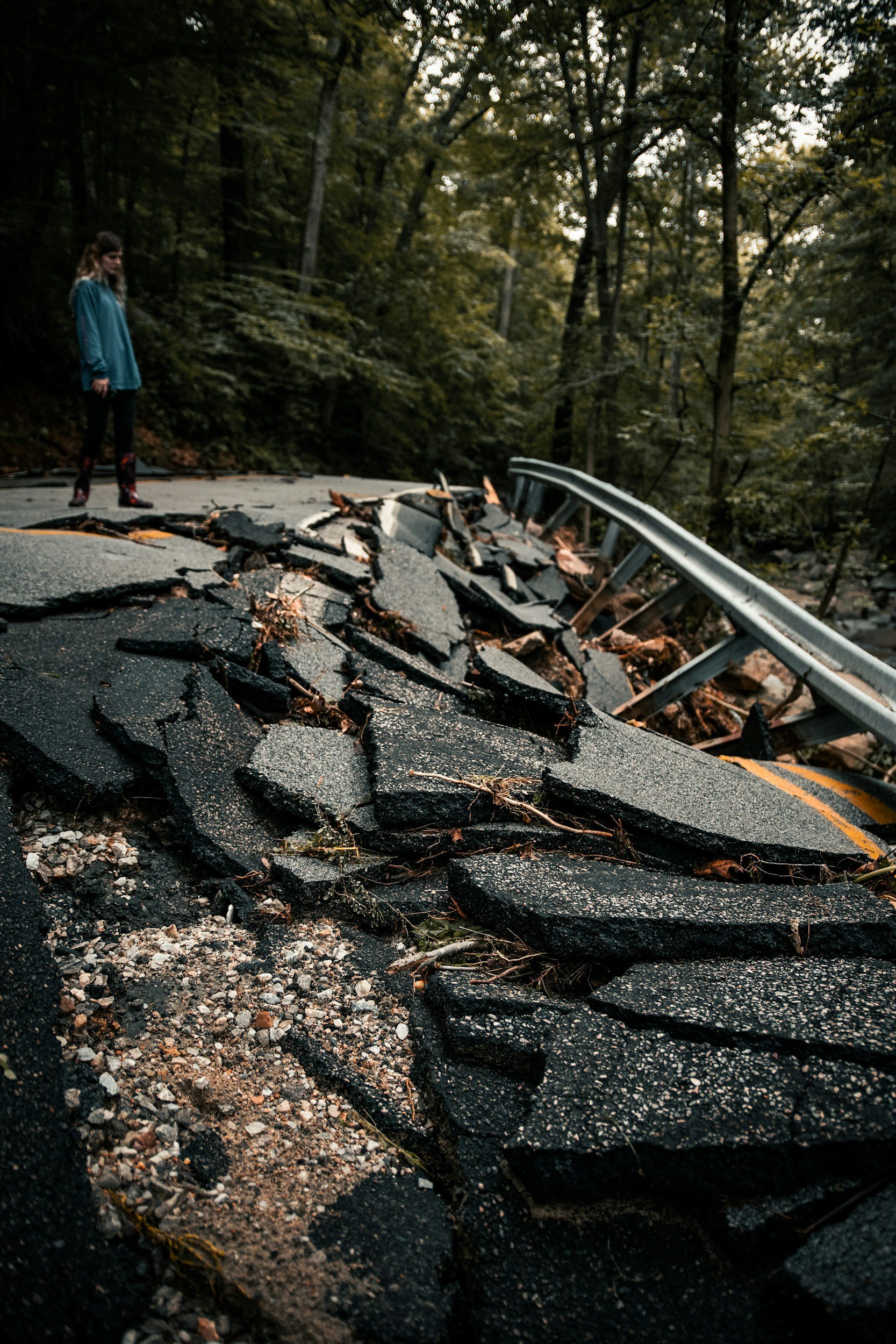 A woman standing on a damaged road with a collapsed section and a metal guardrail, surrounded by dense trees in a forest.