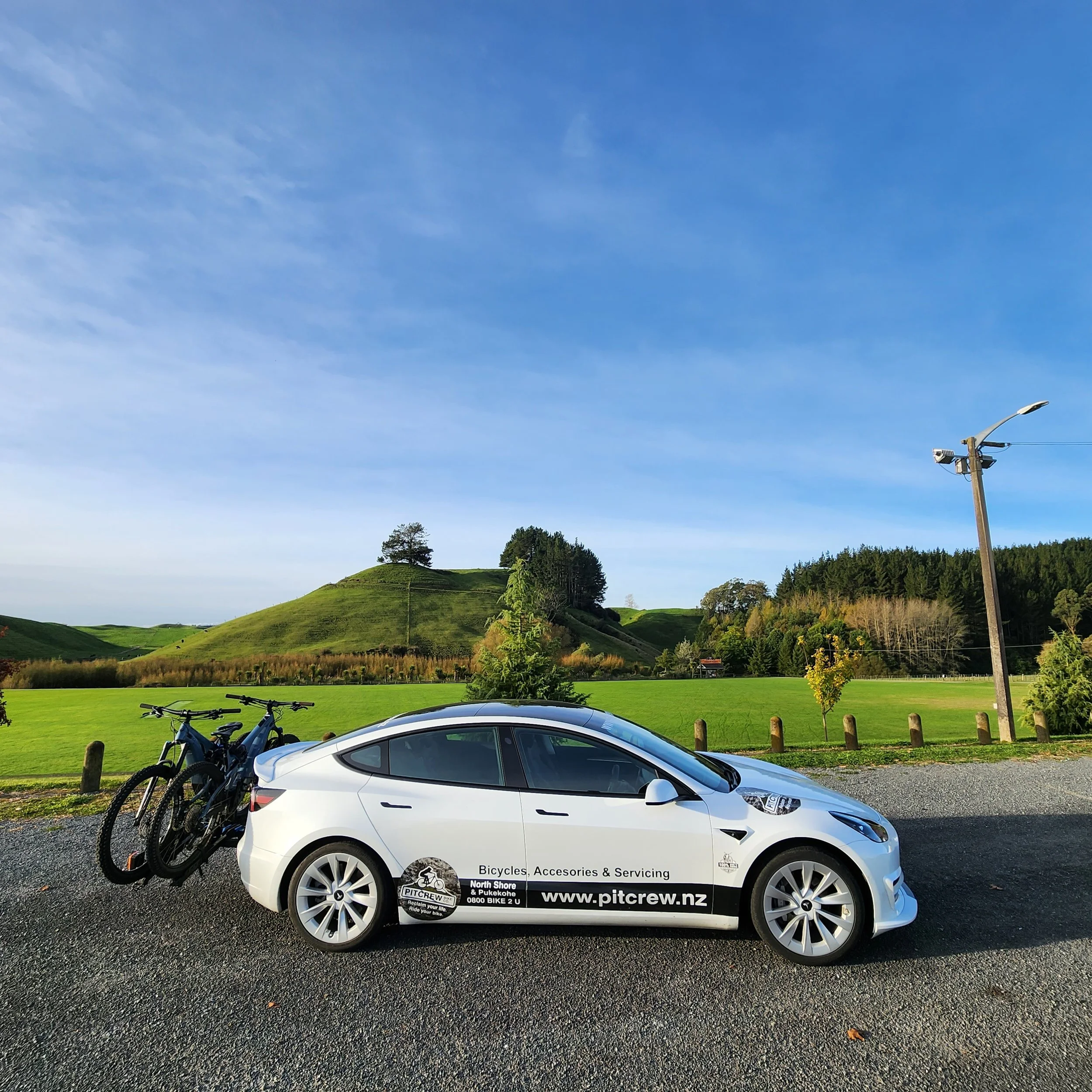 A white electric car with two bicycles mounted on the back, parked on a gravel lot. The car has signage advertising bicycles, accessories, and servicing, with a website www.pitcrew.nz. The background features green hills, trees, and a clear blue sky.