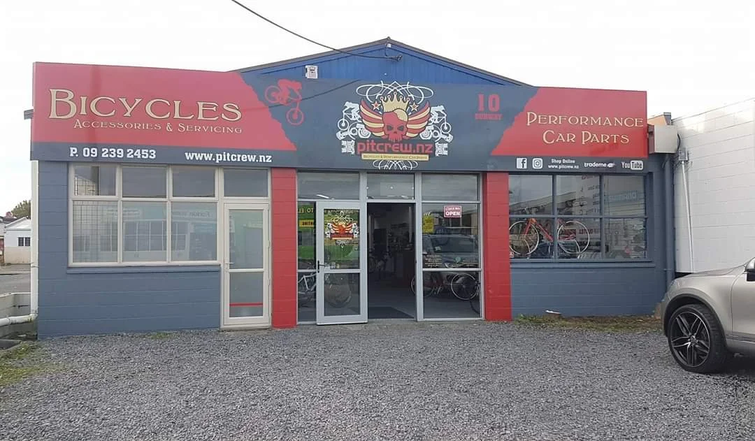Bike shop with red and gray exterior, large windows, and a sign that reads watches and performance car parts, with bikes displayed inside.