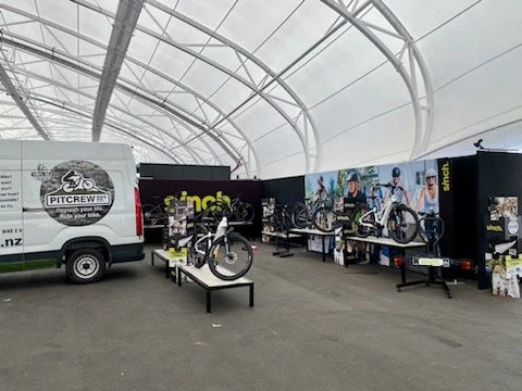 Exhibit of bicycles and cycling gear inside a large, transparent, arched structure. A white van with a Pitcrew logo is parked nearby.