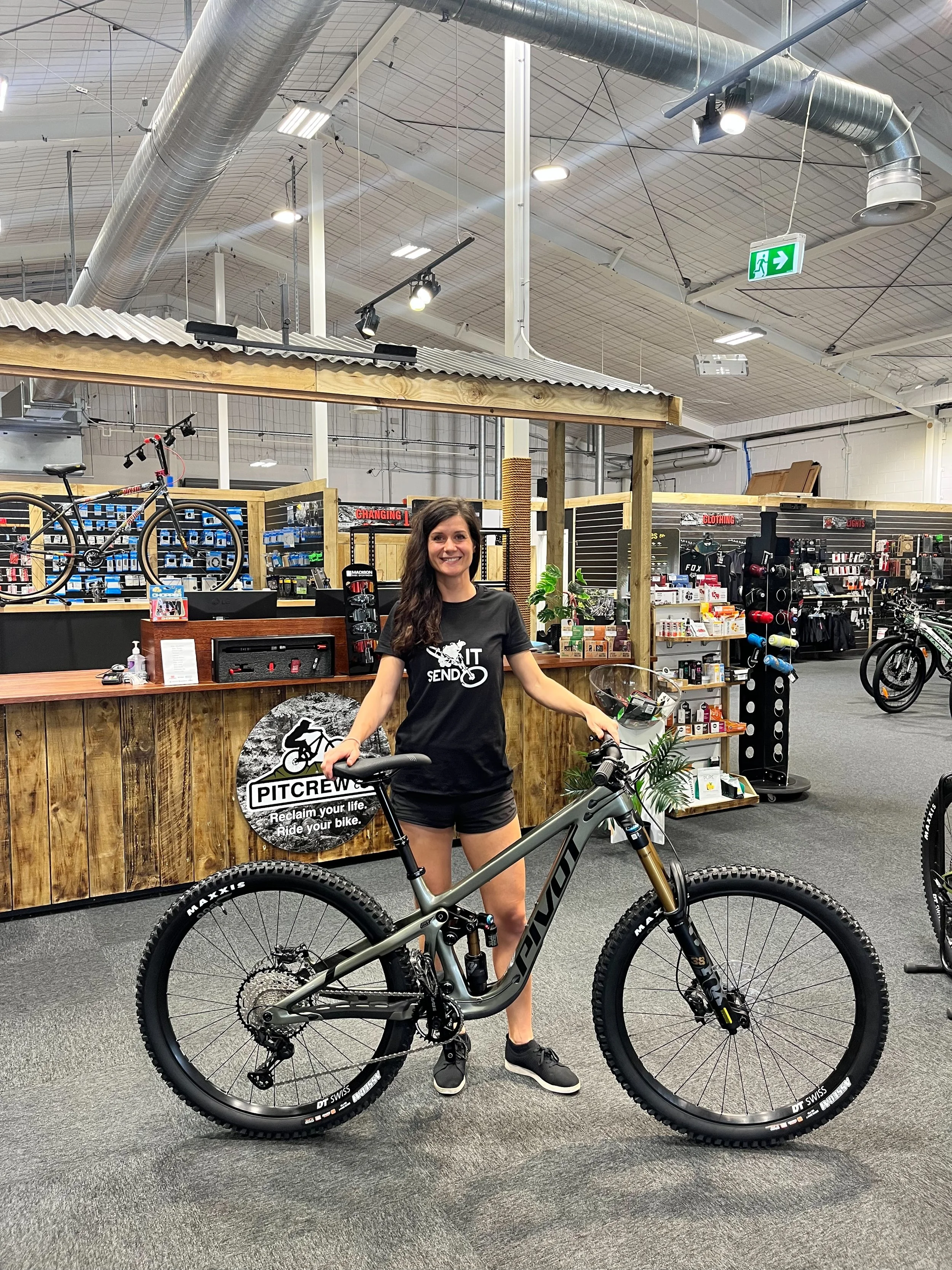 A woman standing inside a bike shop holding a gray Giant mountain bike, smiling at the camera. The background shows bike accessories, a counter with a small plant, and other bicycles displayed in the shop.