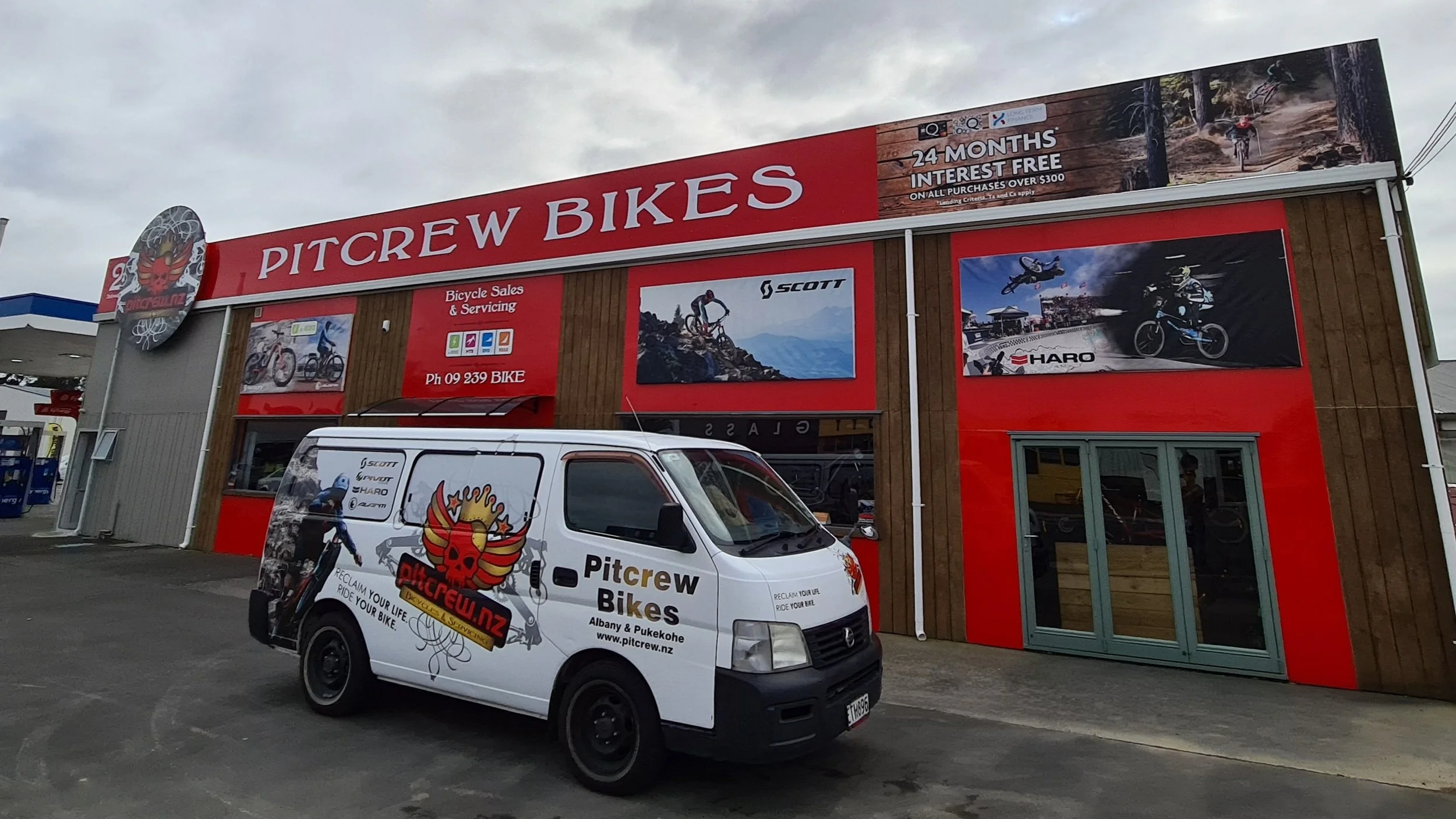 Exterior of a Pitcrew Bikes store with a white delivery van parked in front, featuring logos and graphics of mountain biking brands, and advertisements for bike sales and servicing.
