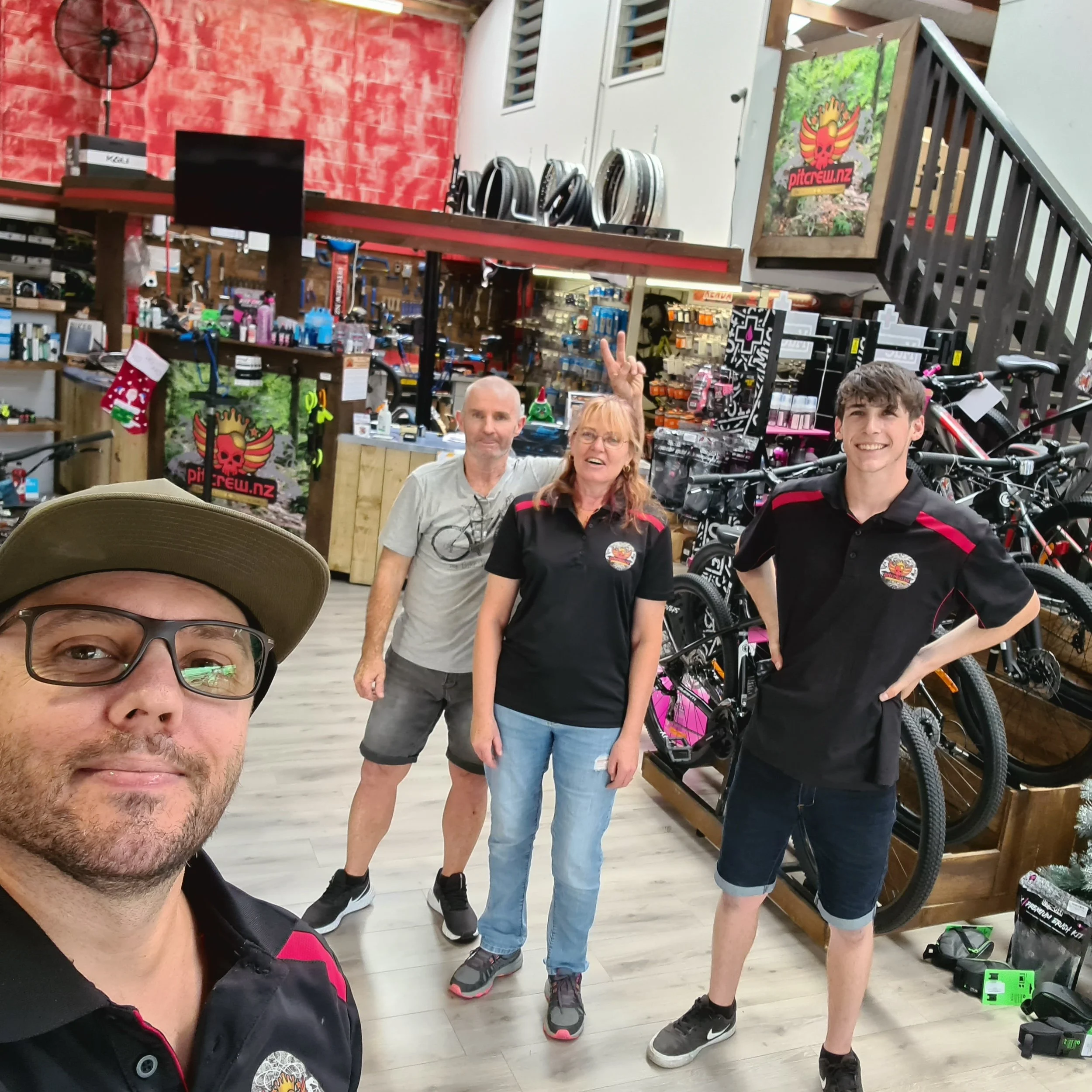 Four people inside a bike shop, three men and one woman, with bicycles and bike accessories displayed behind them. The person in front is taking a selfie. One man is making a peace sign behind the woman.