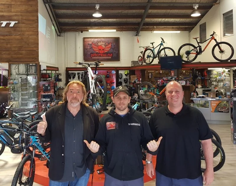 Three men posing inside a bicycle shop, with bicycles and cycling gear displayed in the background.
