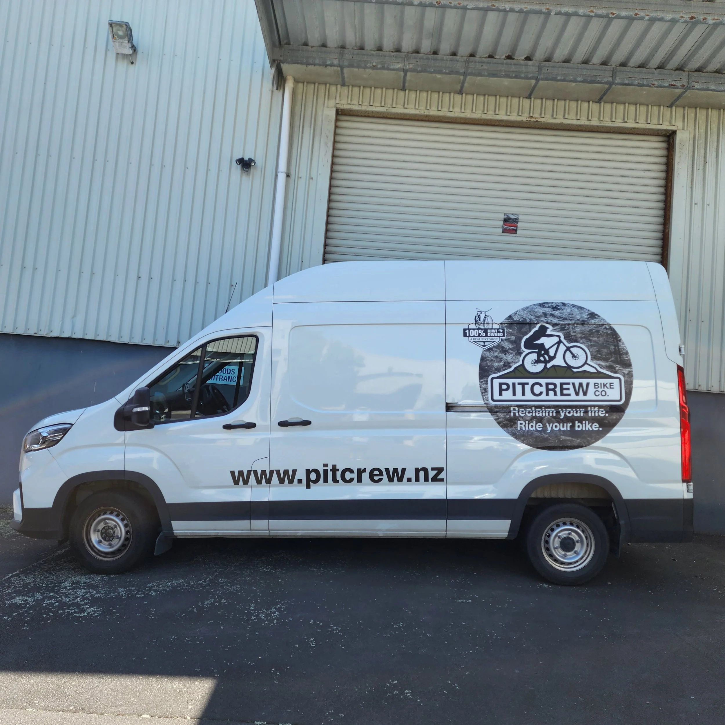 White delivery van with Pitcrew Bike Co. logo and website parked outside an industrial building with a metal roll-up door.