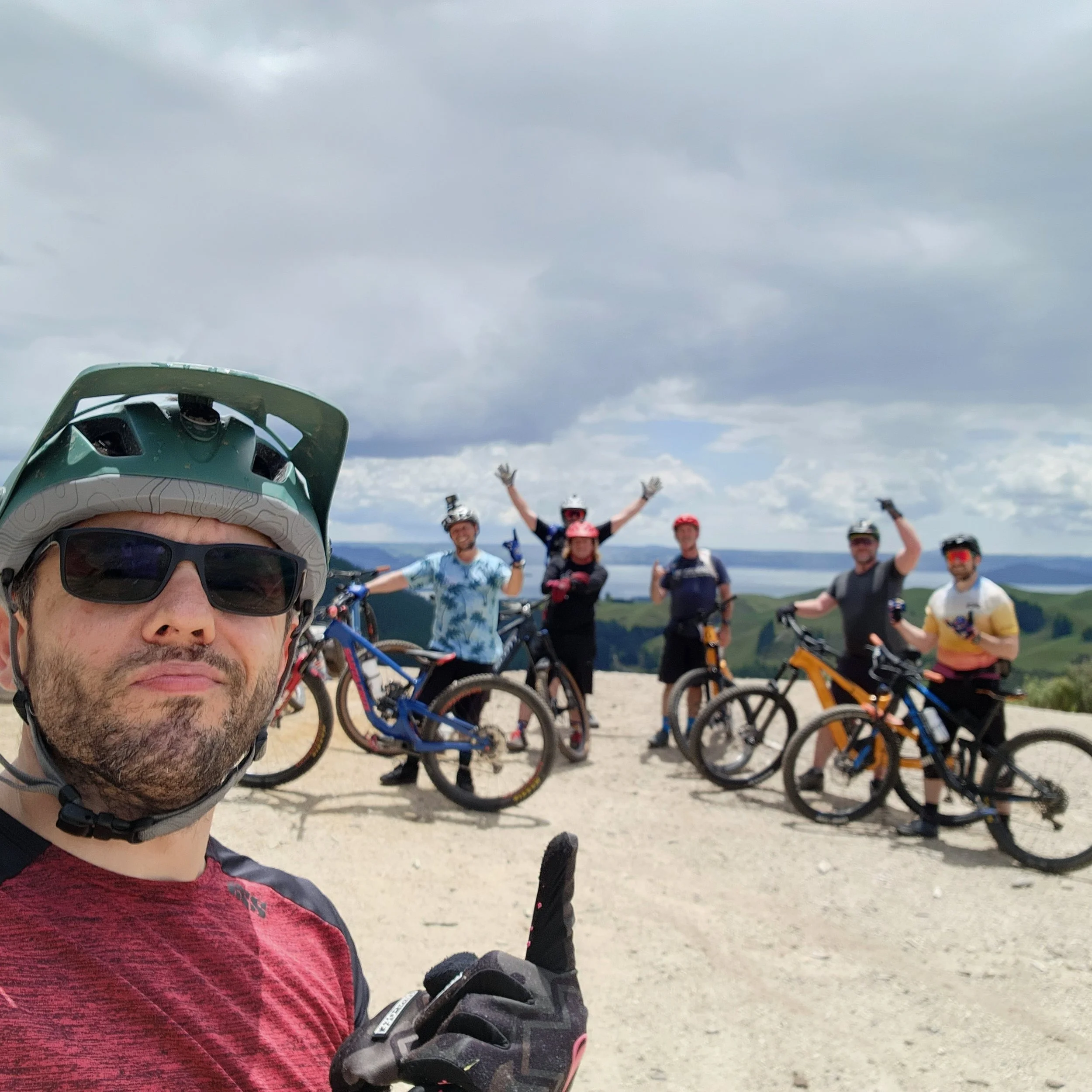 Group of mountain bikers outdoors on a dirt trail with scenic hills and water in the background, one person in the foreground taking a selfie