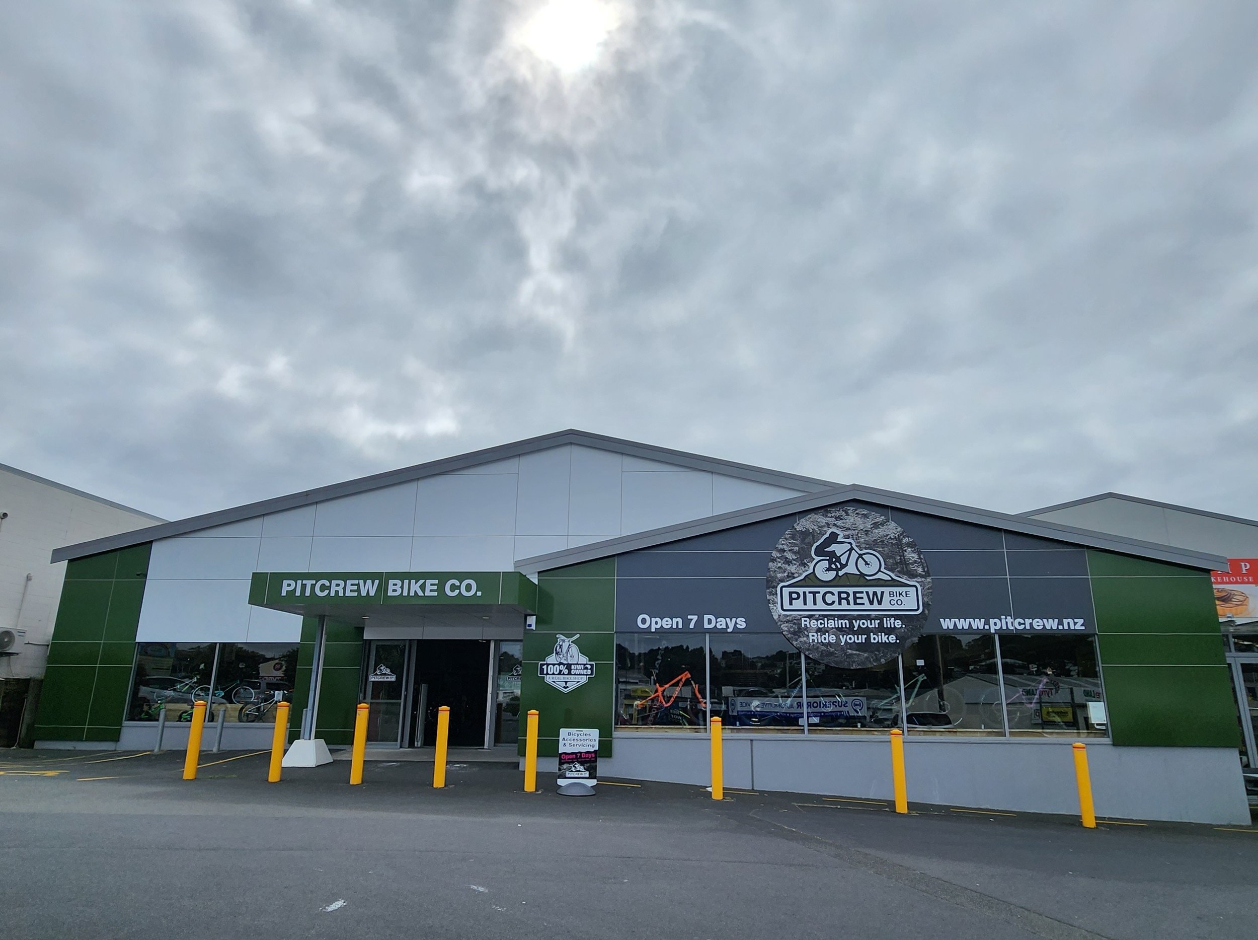 Exterior of Pitcrew Bike Co. store with green and white facade, large logo with a cyclist, and yellow parking barriers in front.