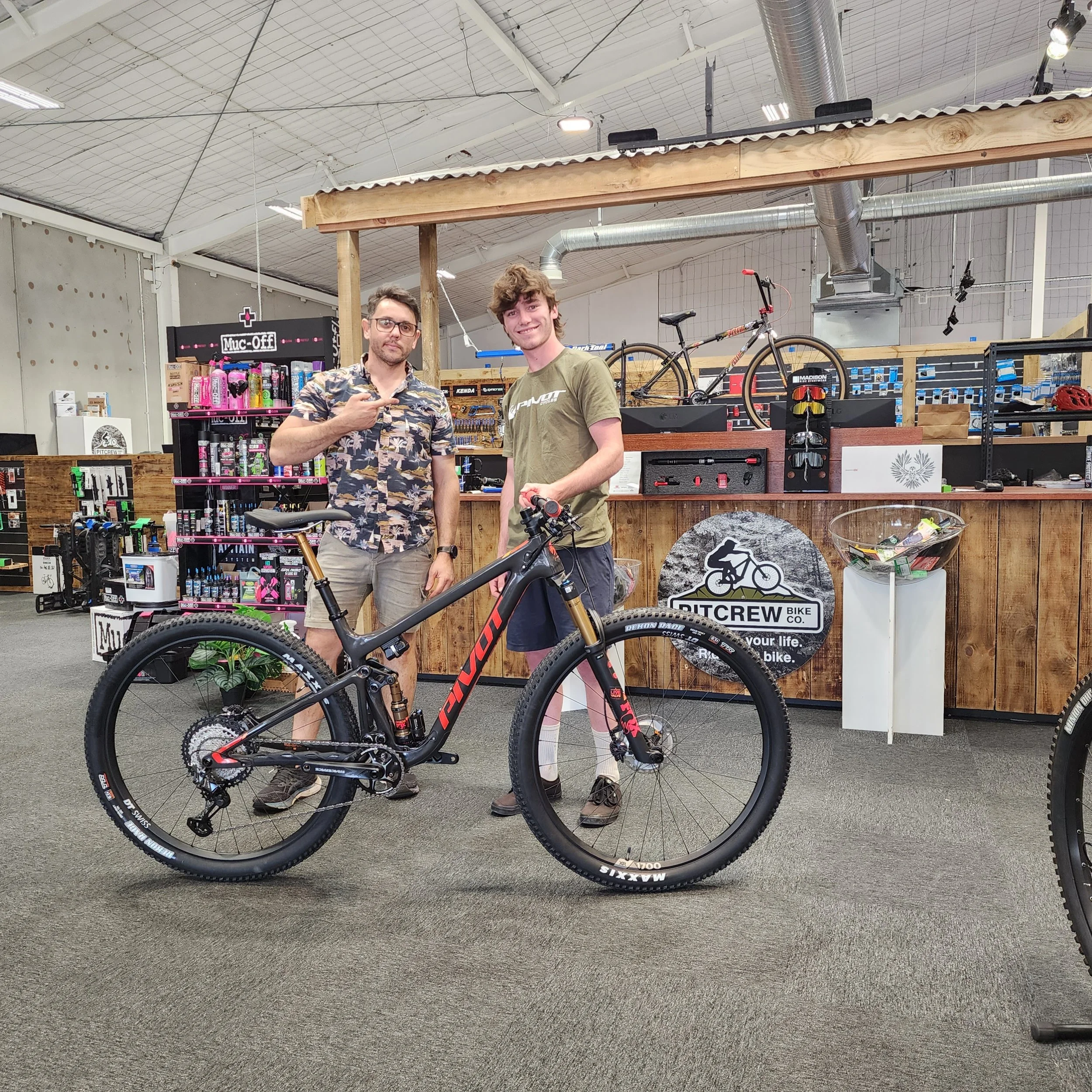 Two men standing beside a black and red mountain bike inside a bike shop, with bike accessories displayed on shelves and a wooden counter in the background.