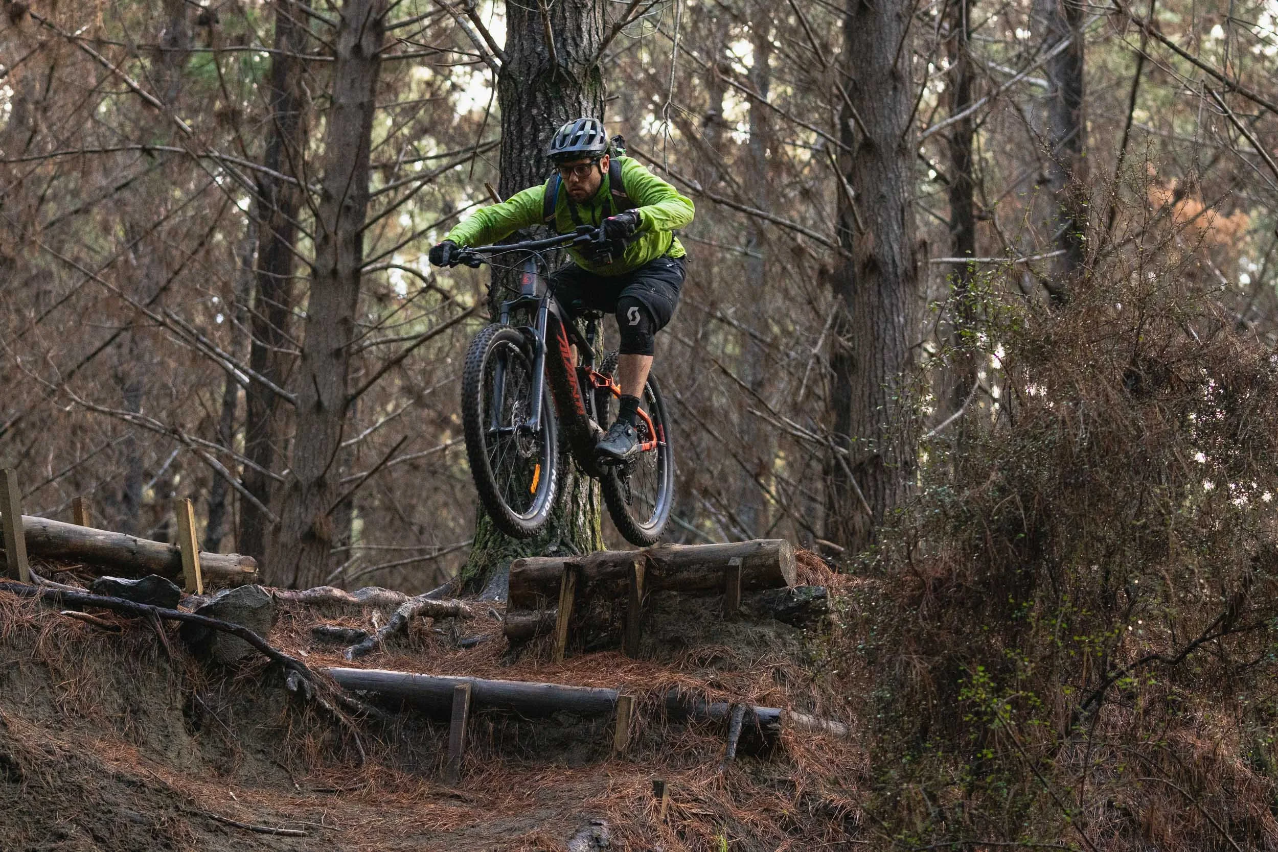 A man mountain biking down a trail in a wooded forest, in mid-air over wooden and dirt jumps.