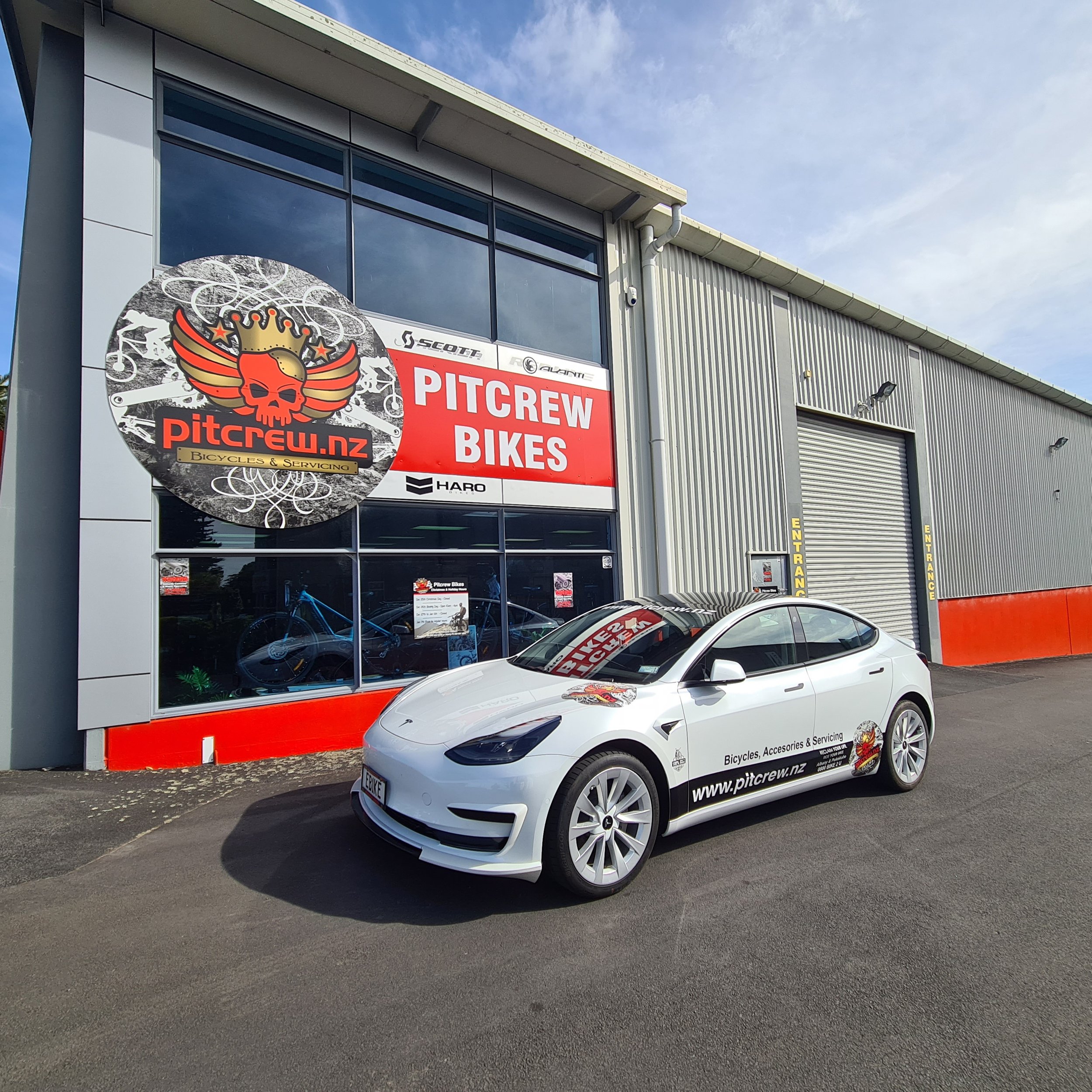 A white Tesla car parked outside a bicycles and servicing shop called Pitcrew BIKES. The shop has a large sign with a skull and wings logo, and the shop's website, pitcrew.nz, is displayed on the side of the car.