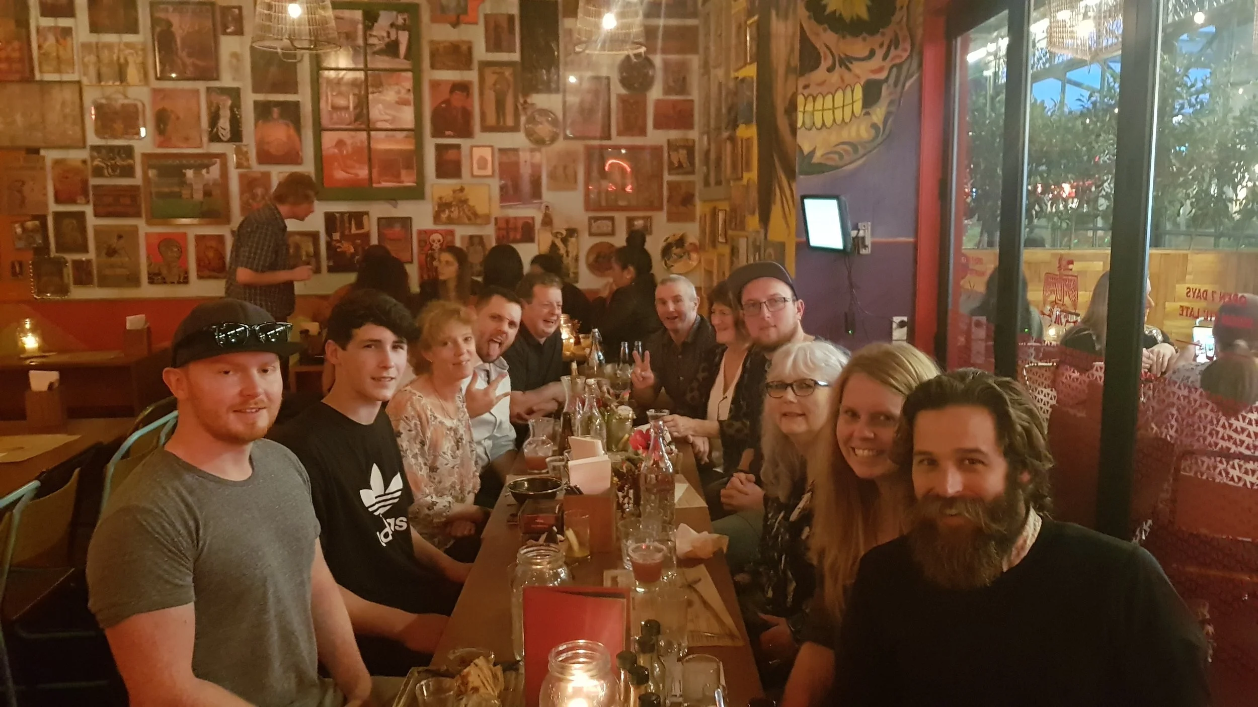 Group of people sitting at a long restaurant table with drinks, smiling and making peace signs, in a colorful and artsy venue with framed pictures and murals on the walls.