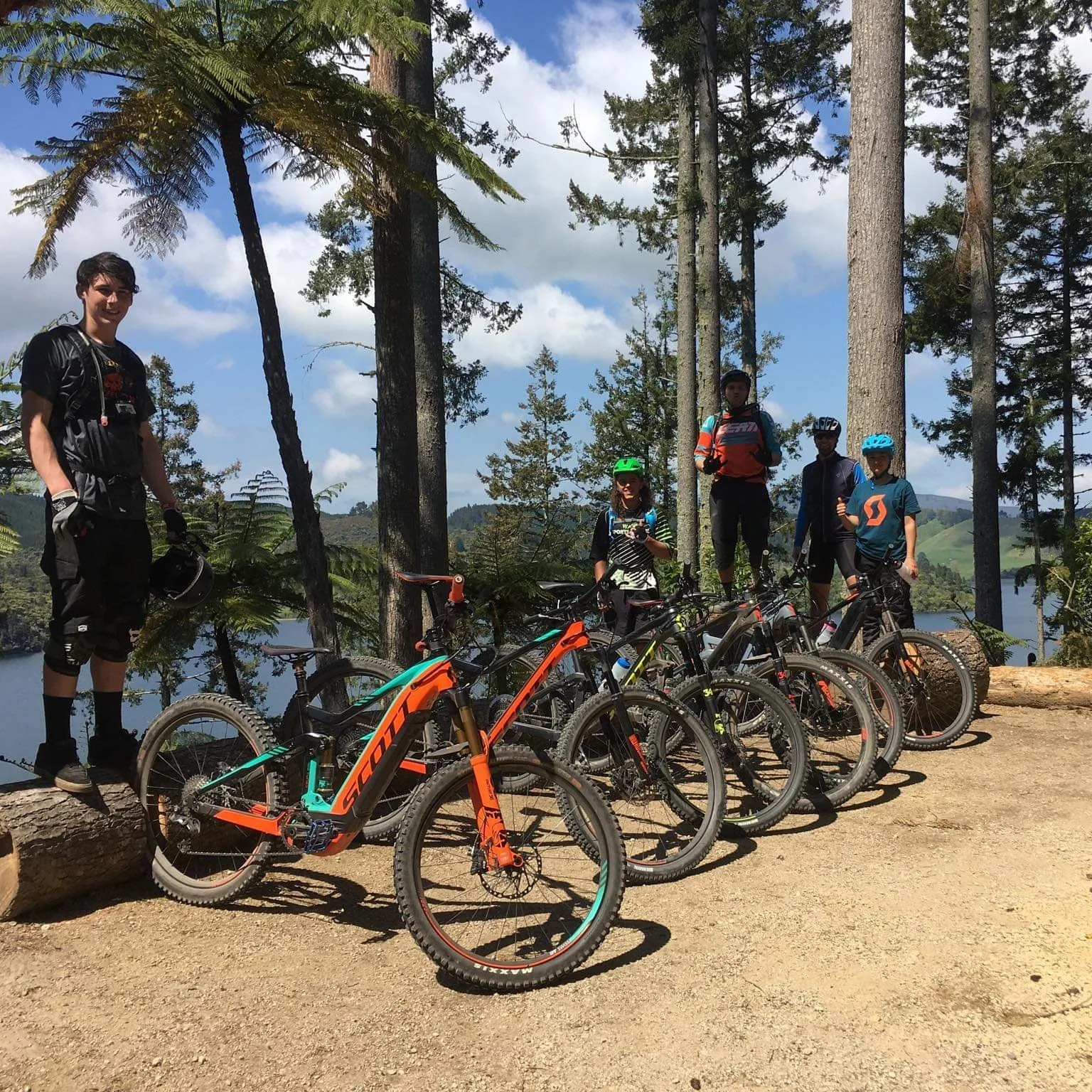 Group of five mountain bikers with helmets, standing and seated with their bikes on a dirt trail near a lake, surrounded by tall trees and hills under partly cloudy sky.