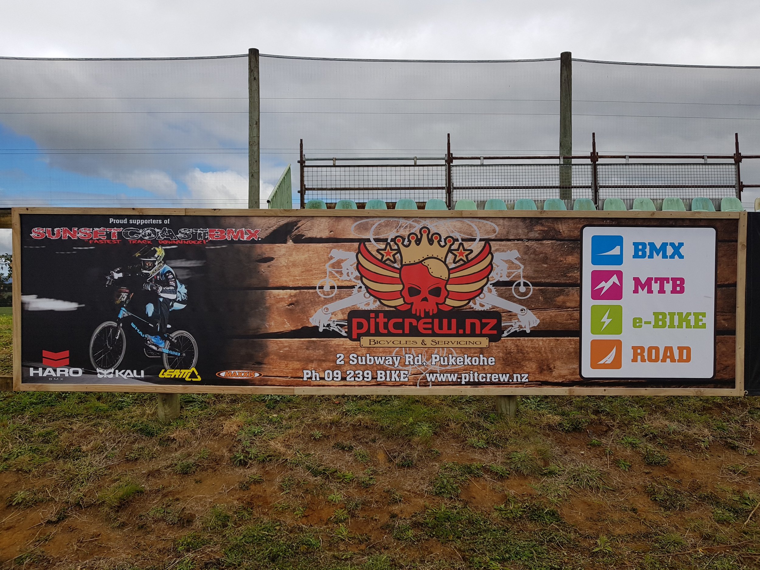 Outdoor banner advertising pitcrew.nz bike shop with a photo of a mountain bike rider, a skull with a crown logo, and a color-coded legend for bike disciplines: BMX, MTB, e-BIKE, and ROAD.