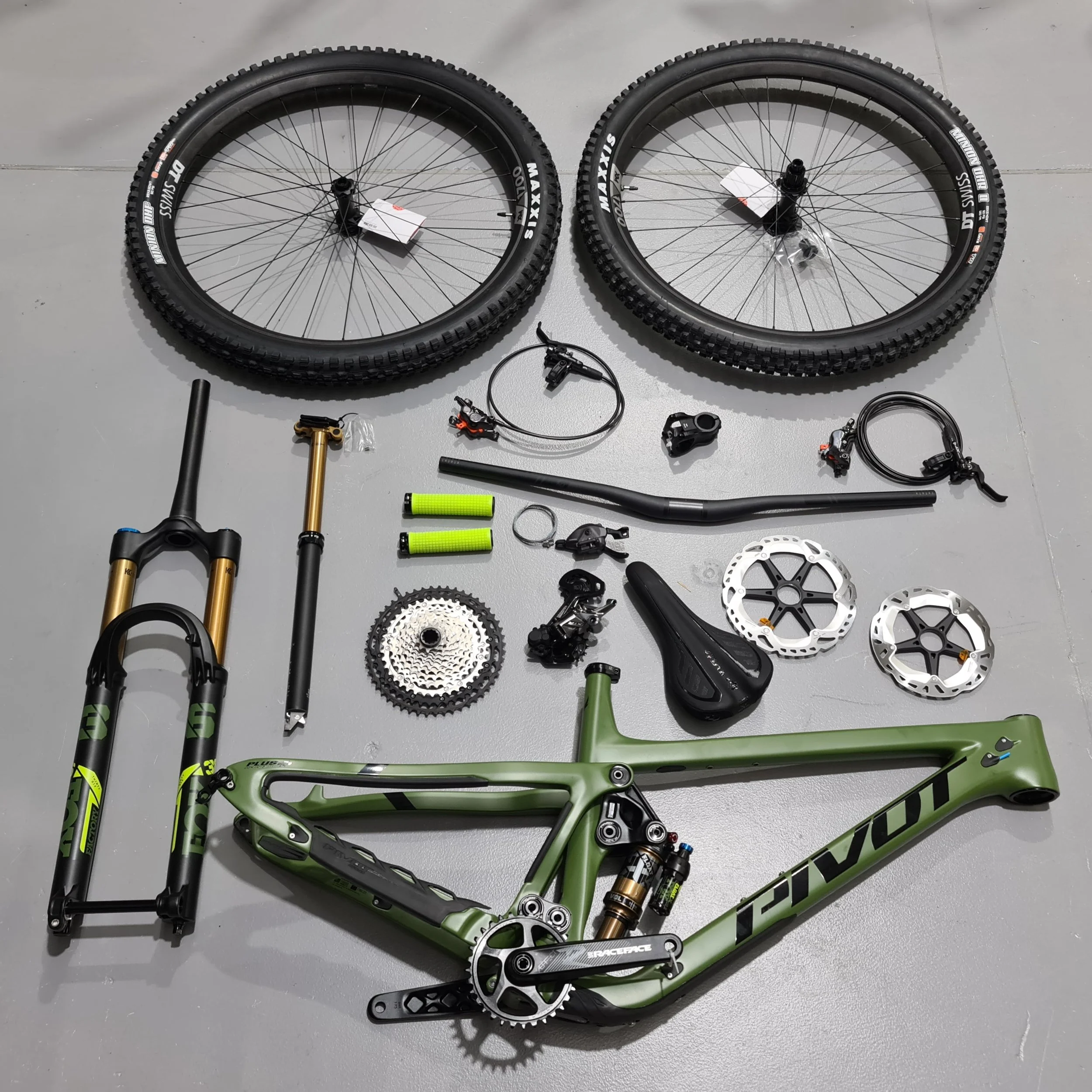 Disassembled green mountain bike with front suspension fork, wheels, crankset, and other components laid out on a gray floor.