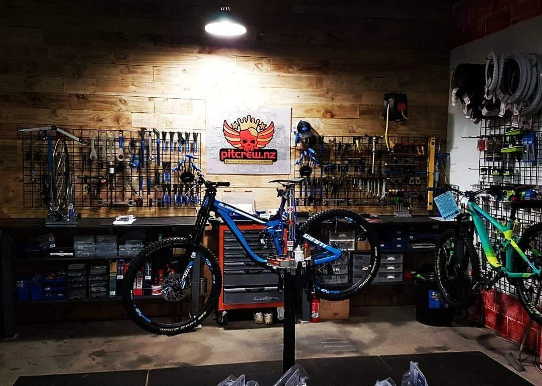 A bike shop with two mountain bikes mounted on a workbench. The wall behind has a wooden panel background with tools organized on pegboards. There is a sign with a skull and wings above the workbench. The shop has overhead lighting and various equipm