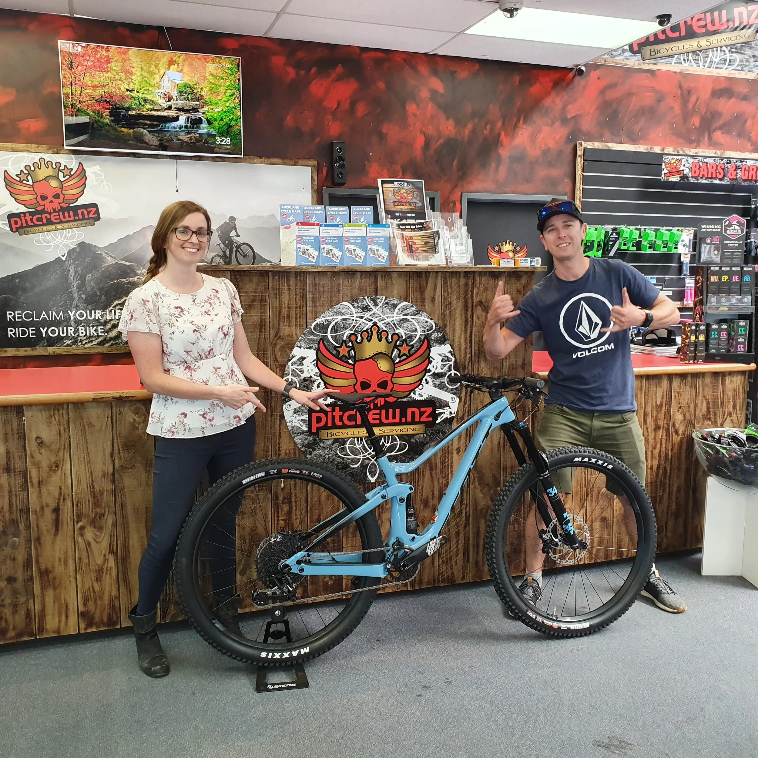 A woman and a man standing behind a wooden bicycle shop counter, with a blue mountain bike in front of them. The woman is pointing at the bike, and the man is making a thumbs-up gesture. The shop's logo features a skull with a crown and wings, and sh