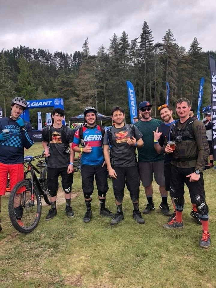Seven men standing outdoors on a grassy field, some wearing mountain biking gear, with a mountain bike, trees, and branded flags in the background.