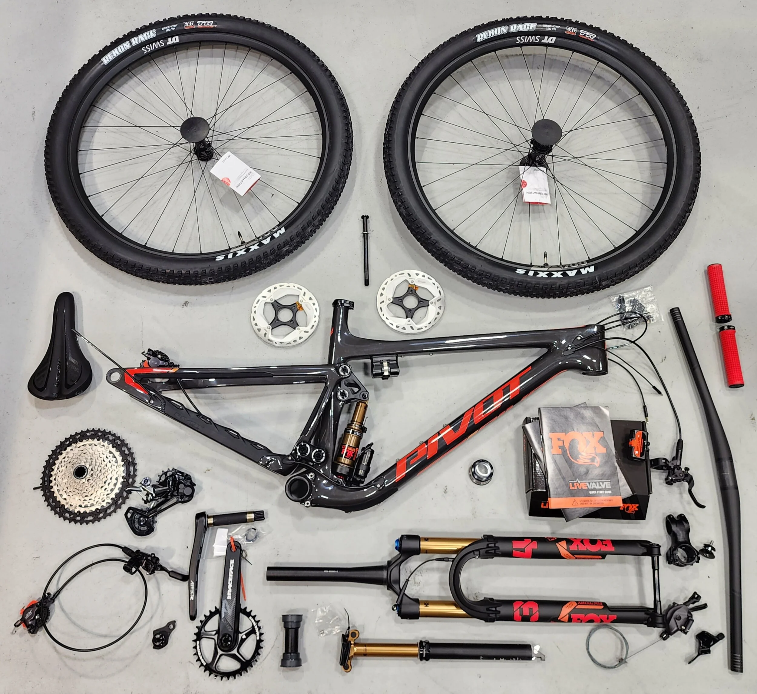 Disassembled mountain bike parts laid out on a gray surface, including two wheels, frame, fork, suspension, pedals, gears, and various small components.