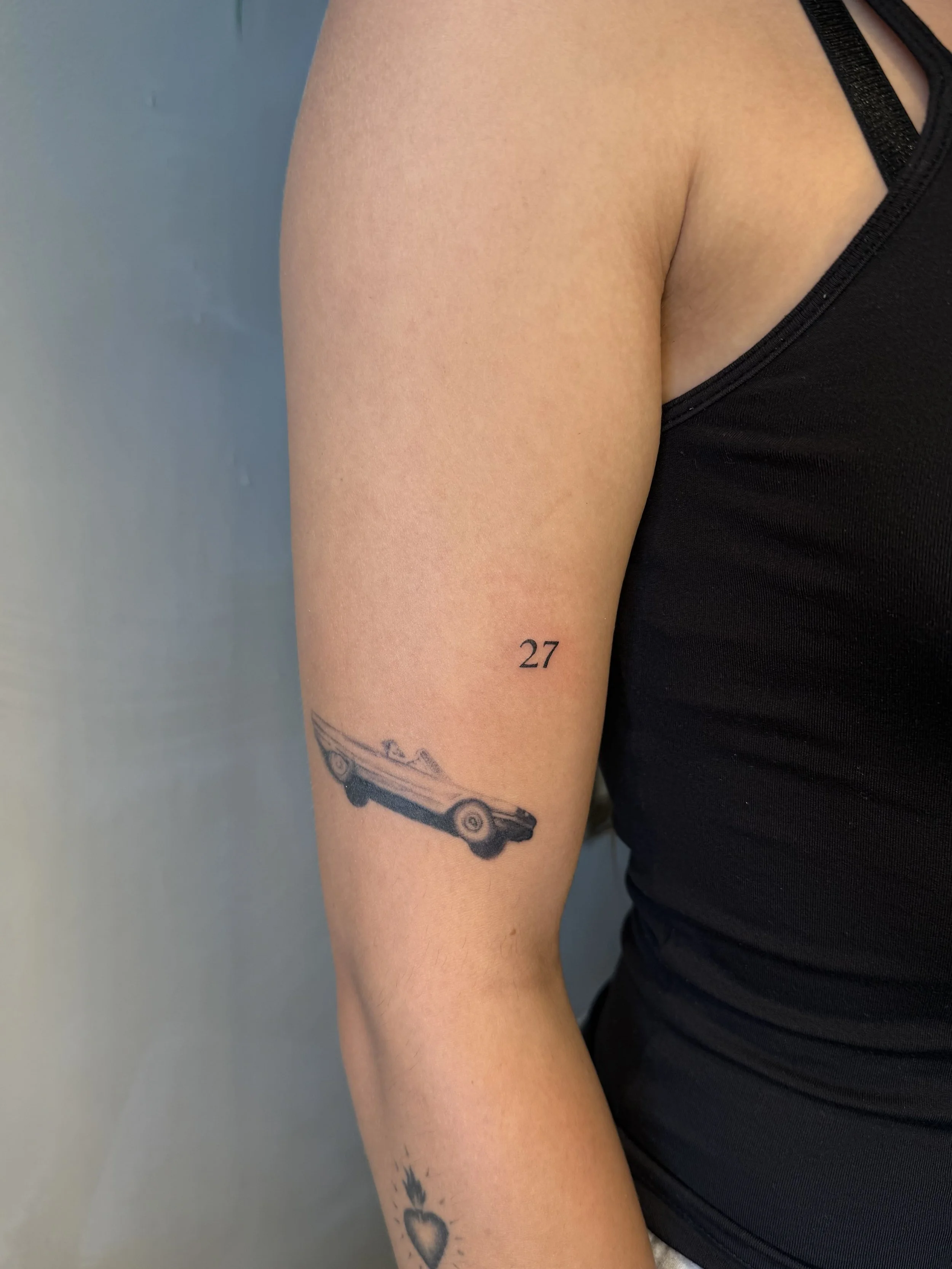 Fine-line vintage car tattoo with small “27” number detail on the upper arm, by Priscilla Shafighi.