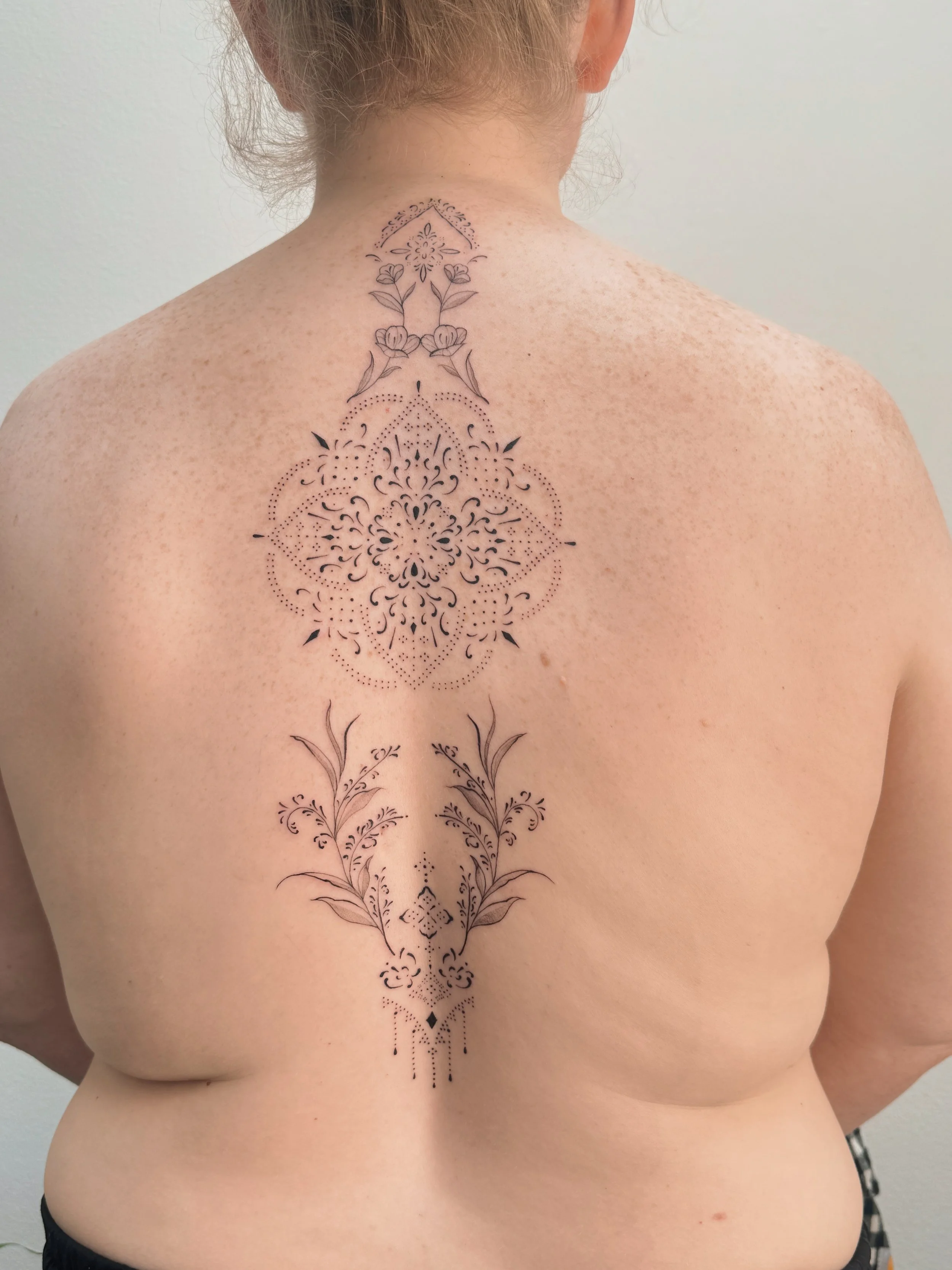 Fine line ornamental back tattoo by Ámya Mila featuring a large central mandala with floral, botanical, and symmetrical decorative elements extending vertically along the spine.