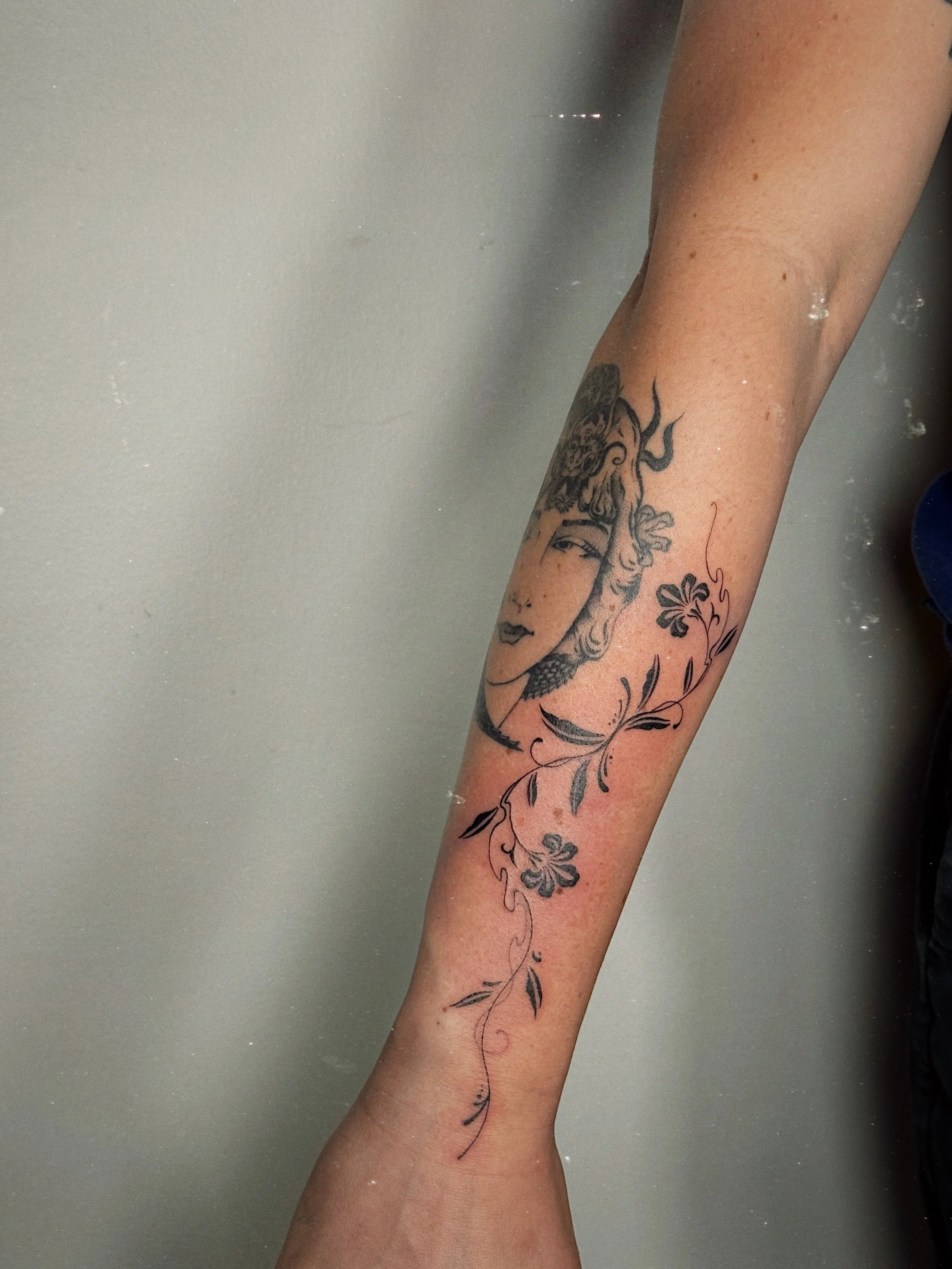 Black and grey fine line tattoo by Ámya Mila featuring a stylized female face with floral and ornamental details flowing along the forearm.