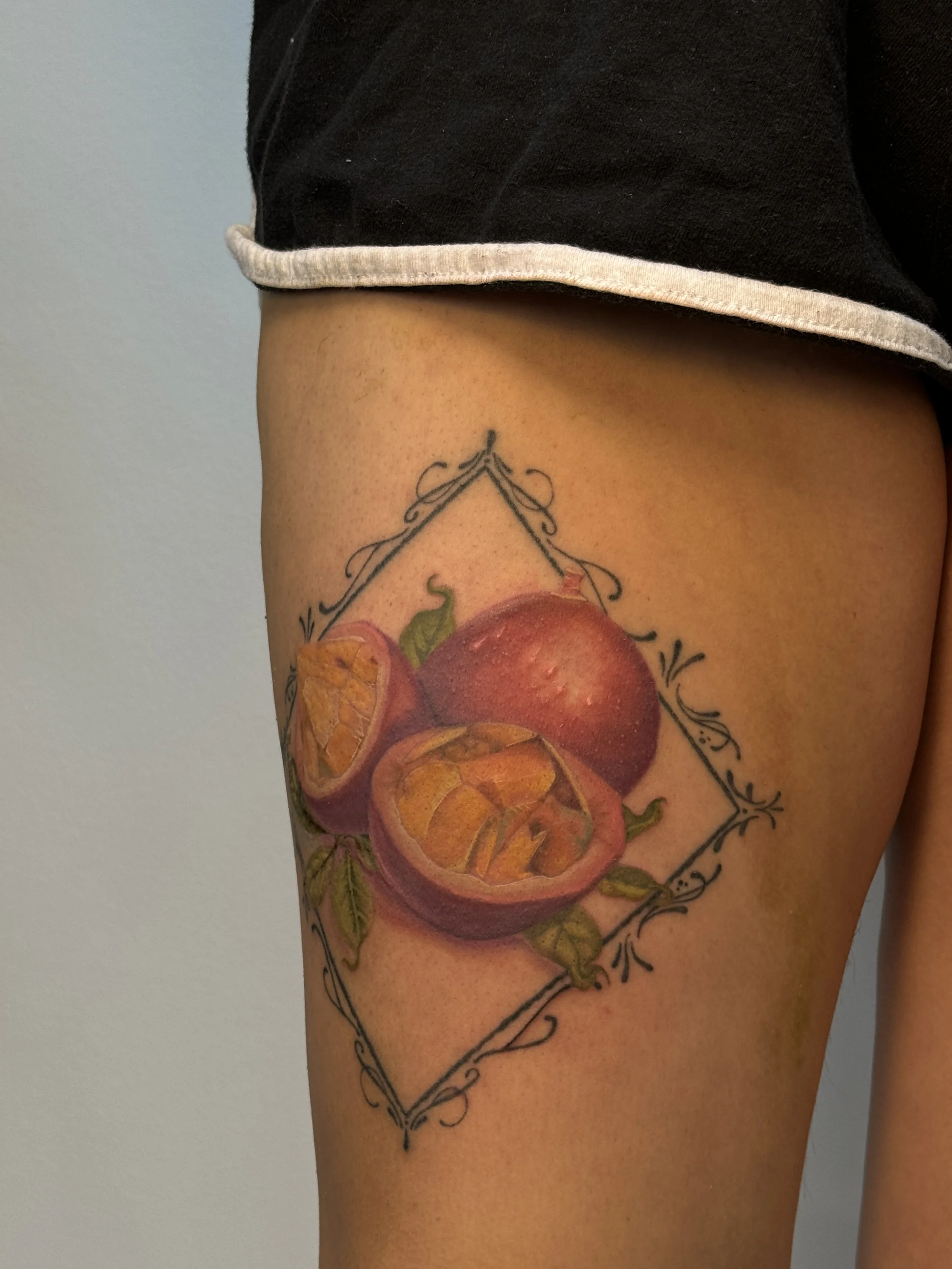 Color passion fruit tattoo by Ámya Mila featuring sliced and whole fruit framed with delicate ornamental linework on the thigh.