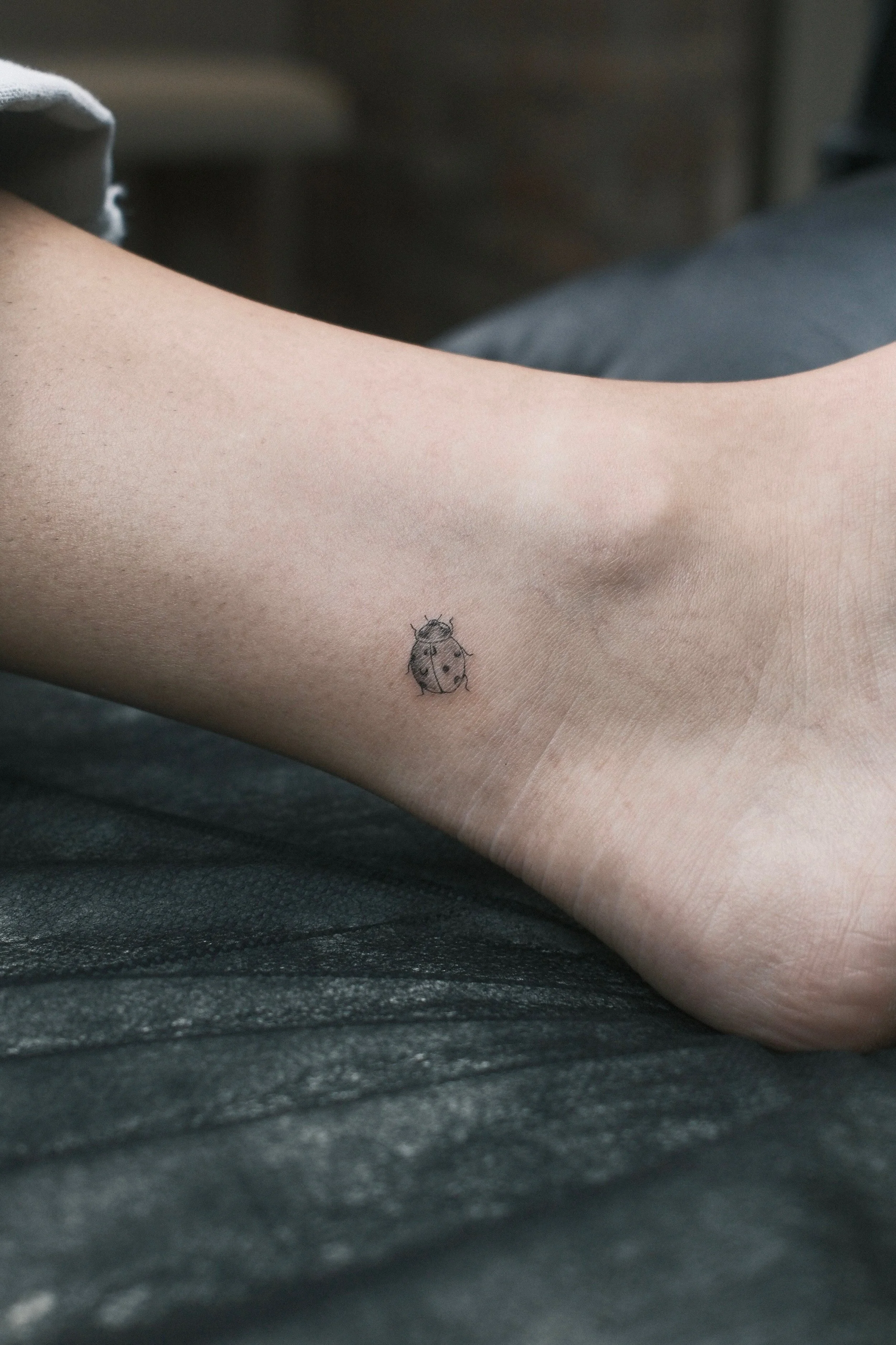 Fine-line ladybug tattoo with delicate black-and-grey shading on the ankle, by Priscilla Shafighi.