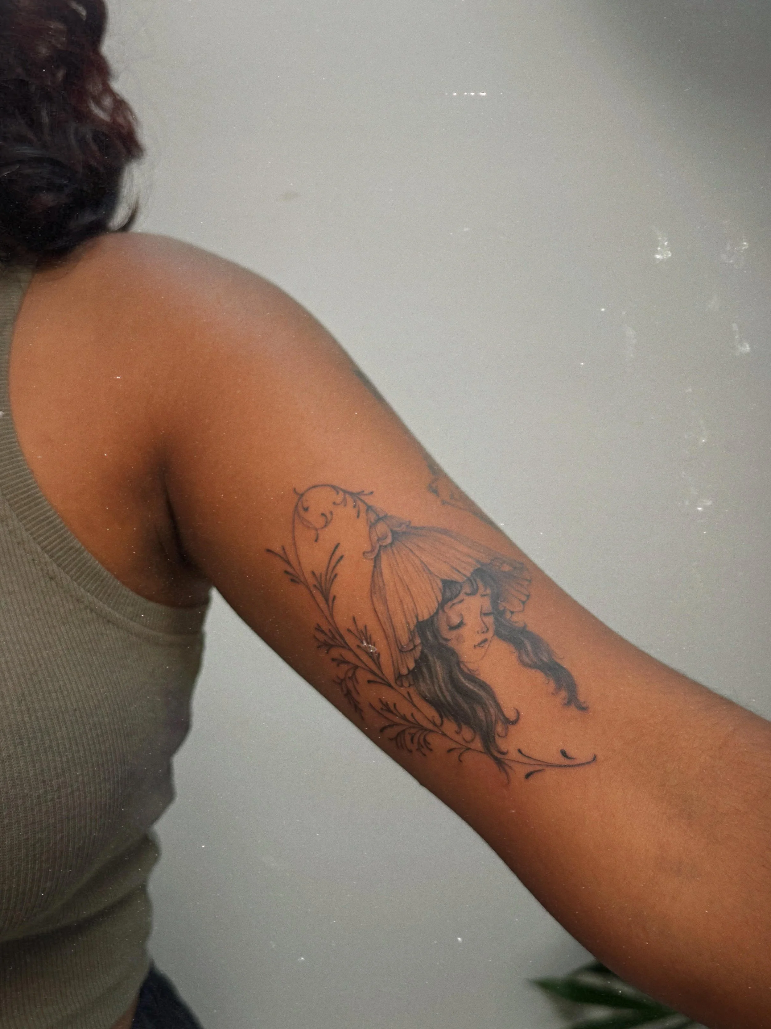 Fine line ornamental tattoo by Ámya Mila featuring an illustrated female figure framed by flowing botanical details on the upper arm.