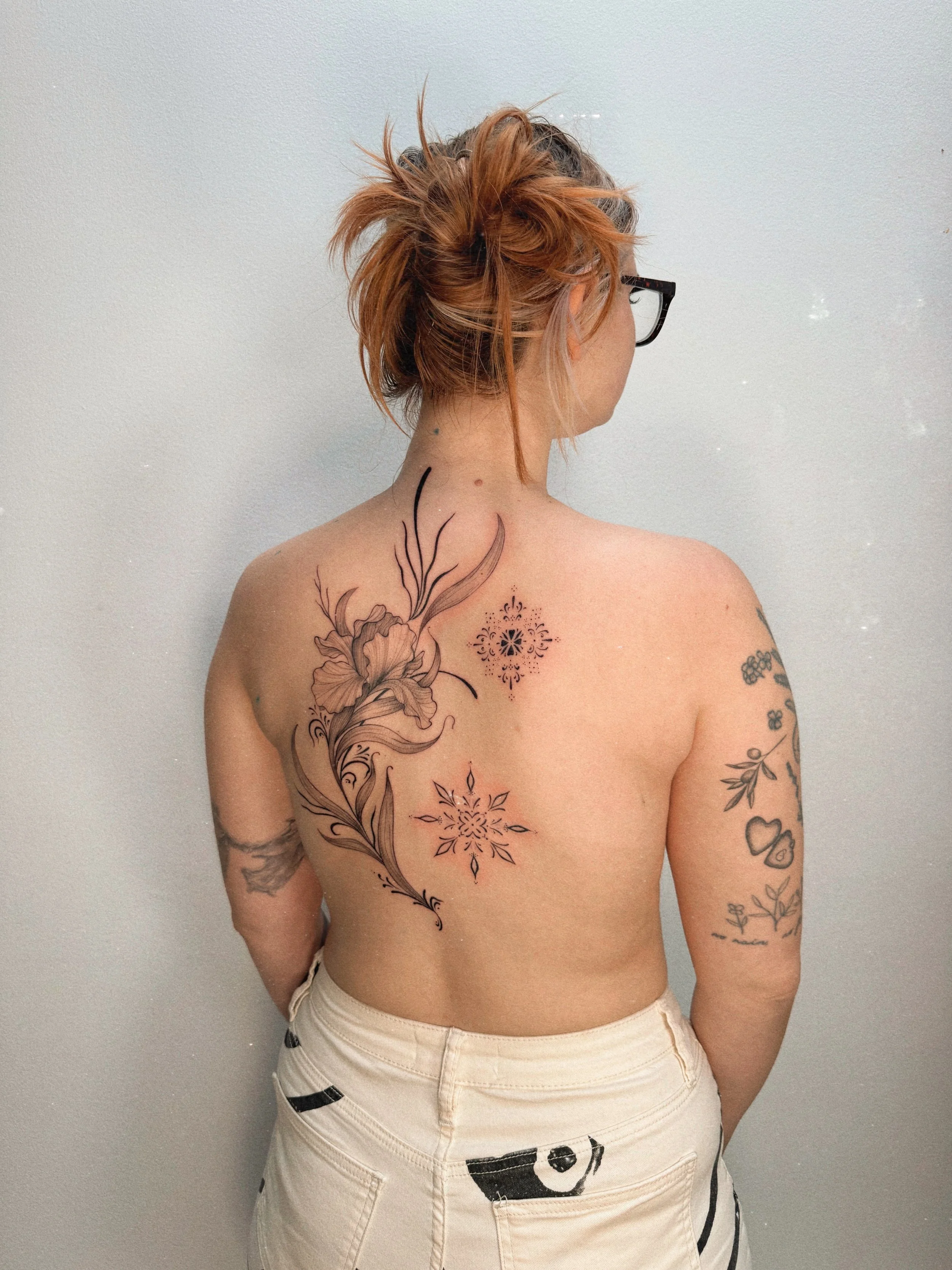 Black and grey floral back tattoo by Ámya Mila featuring large botanical elements and flowing ornamental linework across the upper back.