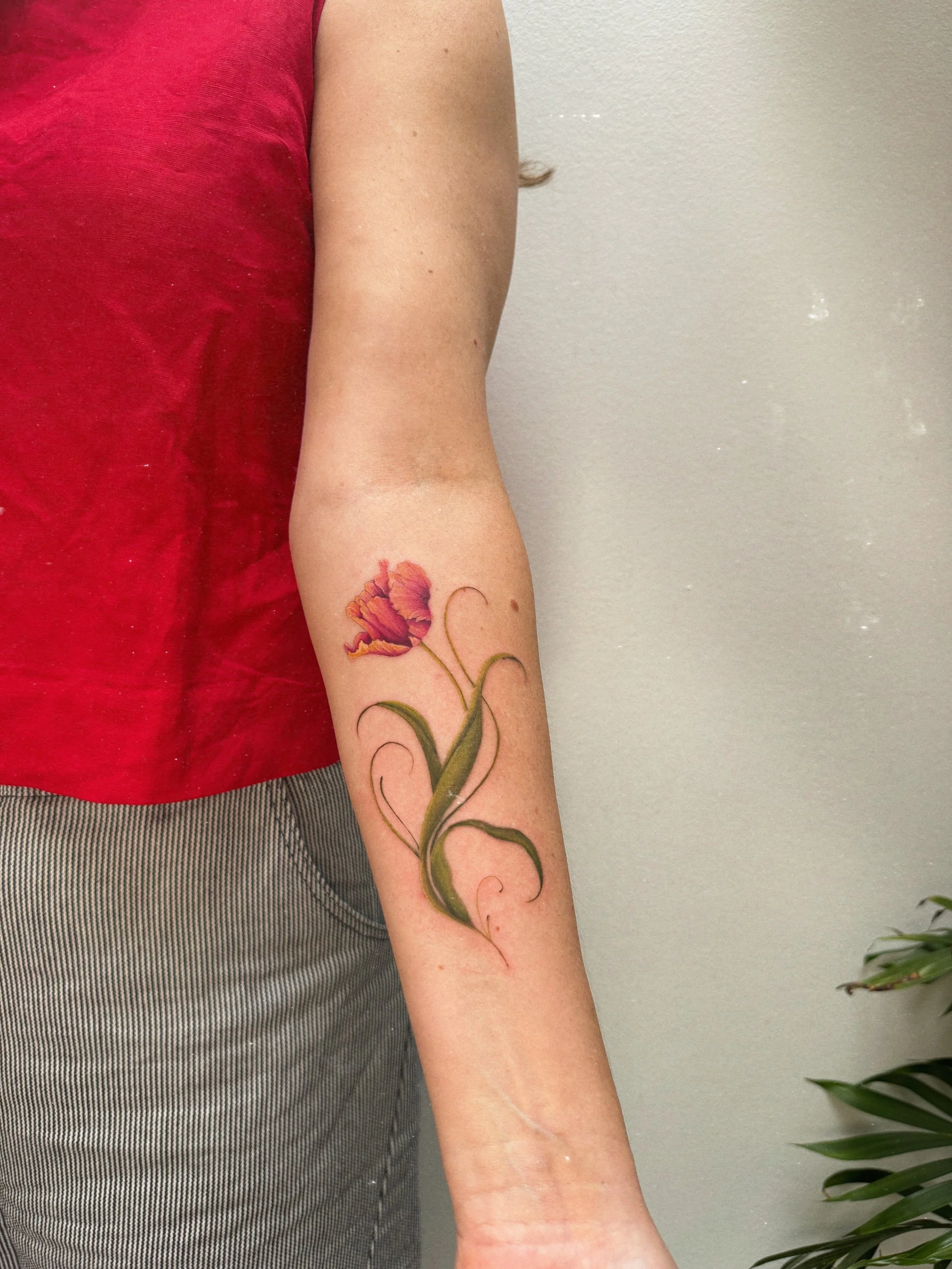Color floral tattoo by Ámya Mila featuring a pink flower with flowing green stems and ornamental linework on the forearm.