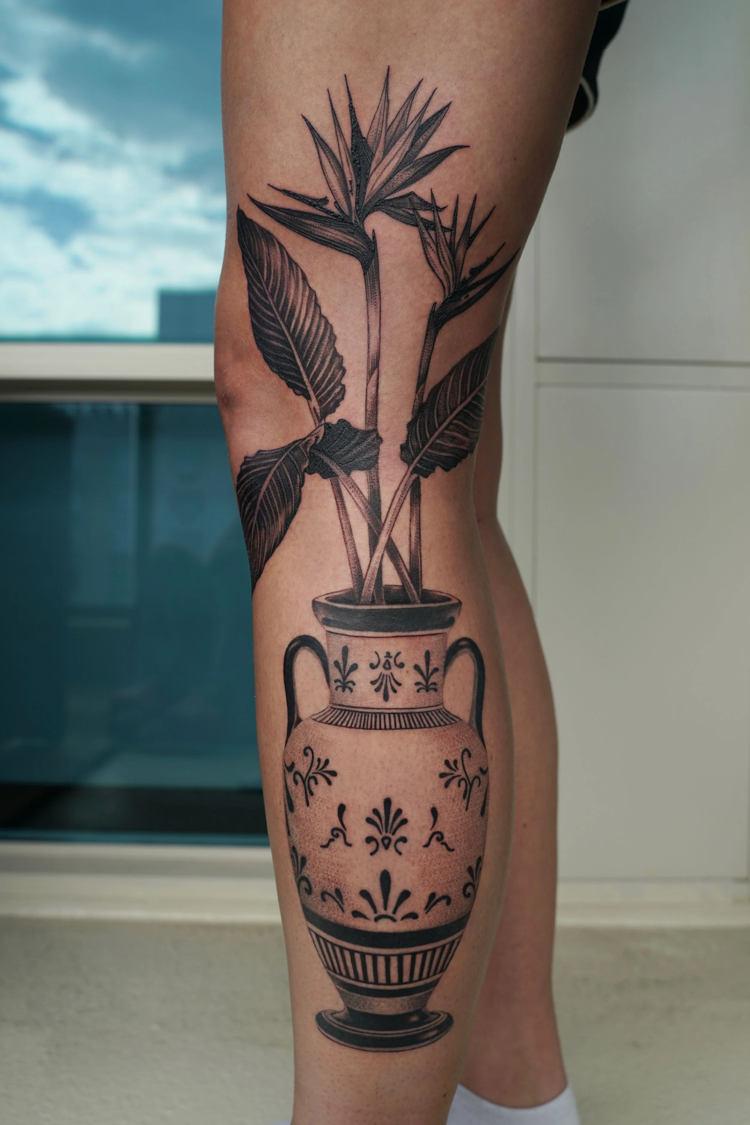 Black-and-grey bird of paradise floral tattoo arranged in a decorative ceramic vase, by Bianca Evangeline.
