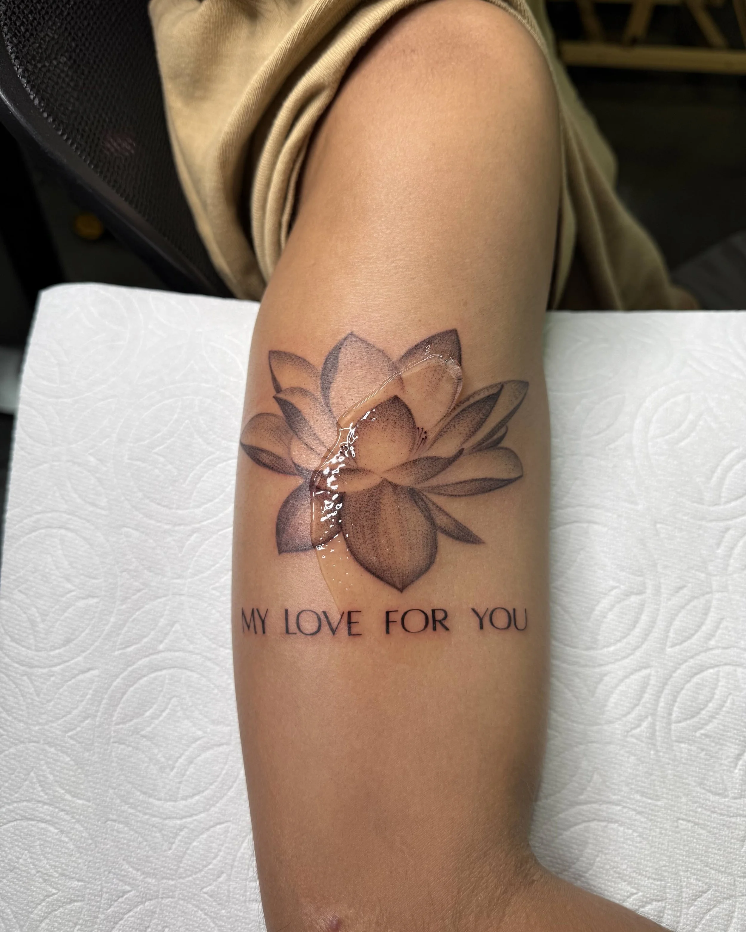 Black-and-grey lotus flower tattoo with “MY LOVE FOR YOU” lettering on the upper arm, by Bianca Evangeline.