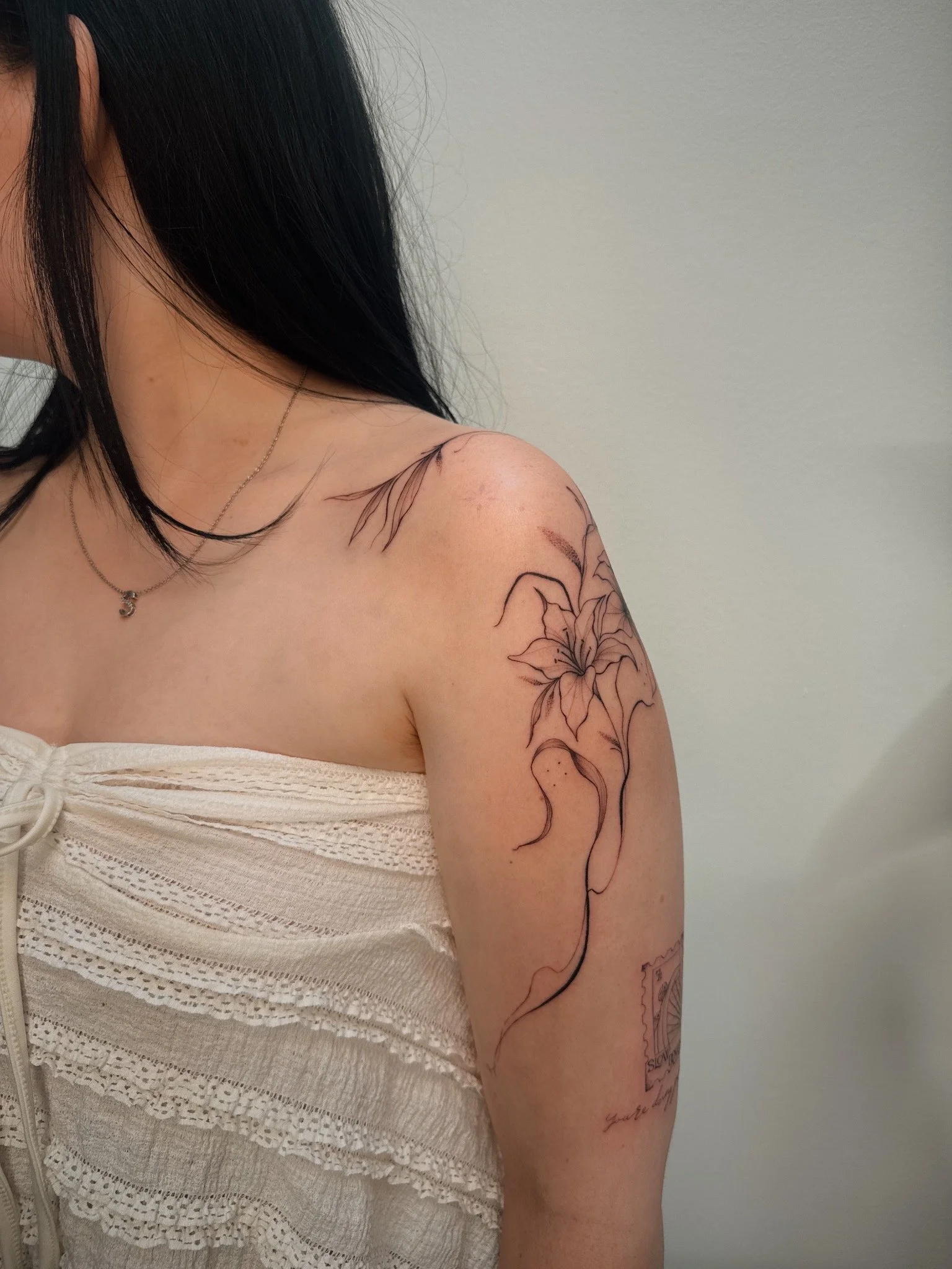 Fine line floral shoulder tattoo by Ámya Mila featuring a large lily with flowing botanical linework extending across the upper arm and collarbone.