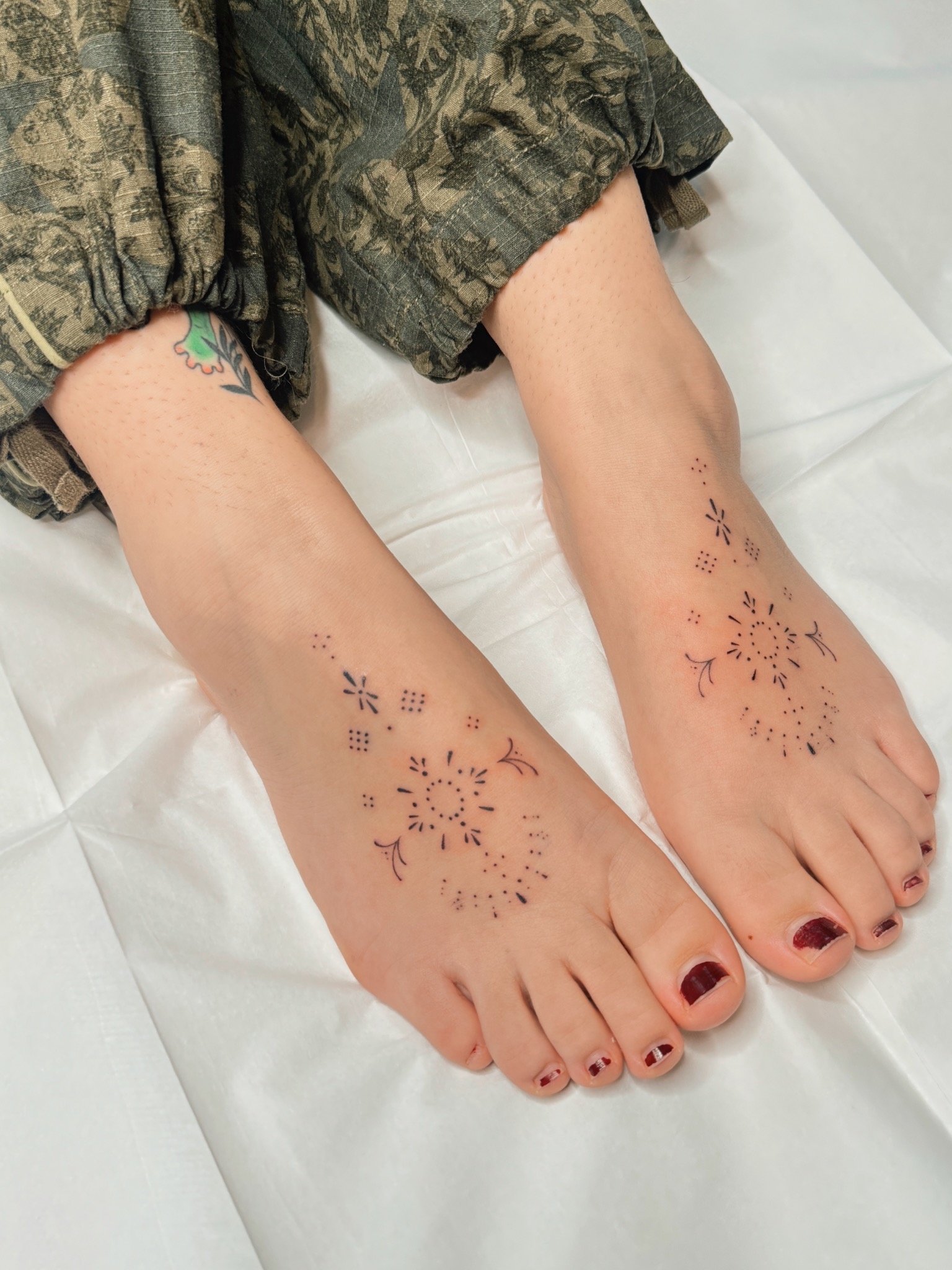 Matching fine line ornamental foot tattoos by Ámya Mila featuring symmetrical dotwork, floral motifs, and delicate decorative details across the tops of both feet.