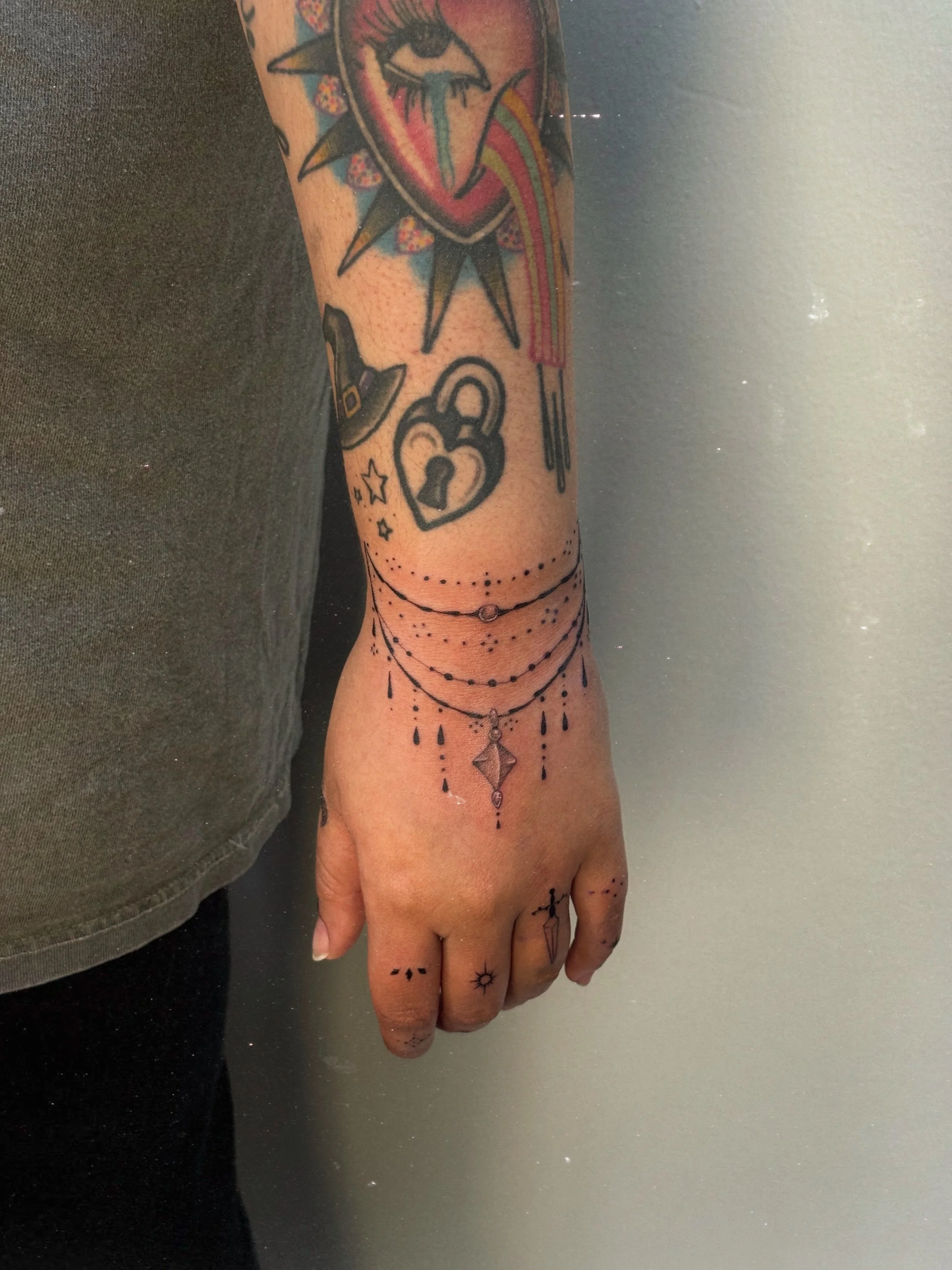 Fine line ornamental wrist and hand tattoo by Ámya Mila featuring layered jewelry-inspired chains, dotwork, and a central hanging pendant detail.