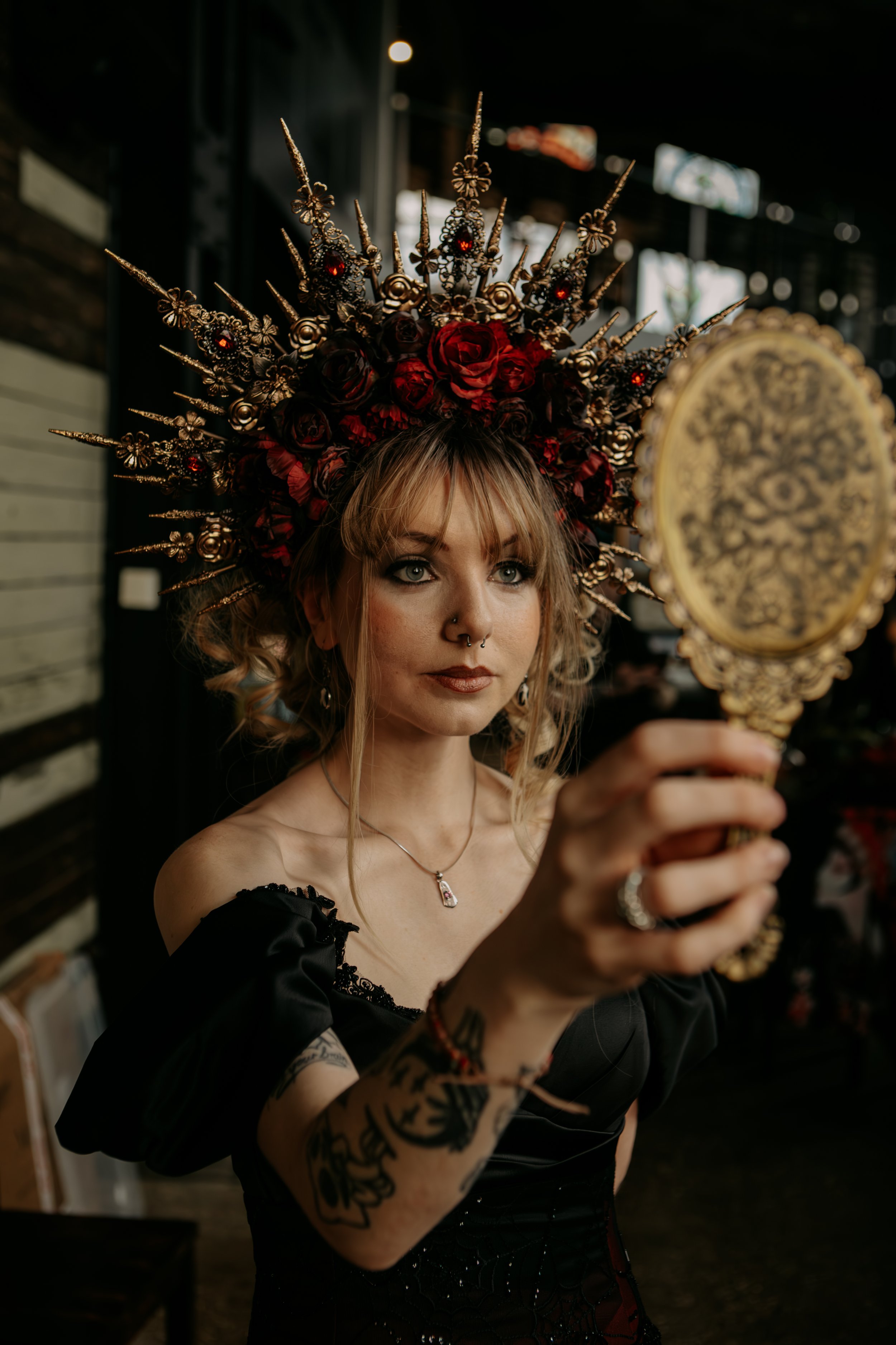 Woman with blonde hair wearing an ornate headpiece made of red roses and gold spikes, holding a vintage handheld mirror, in a dimly lit indoor setting.