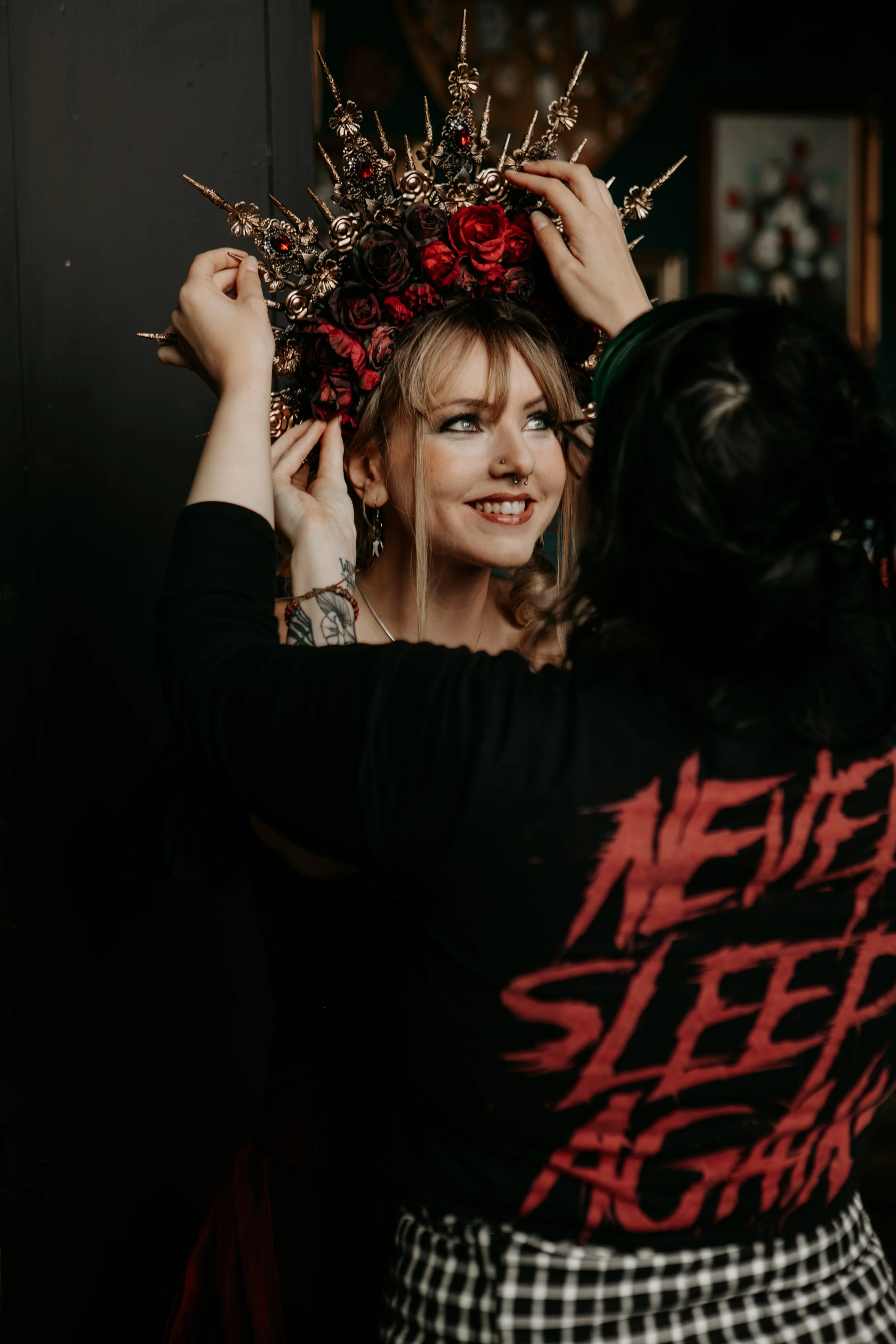 A woman with makeup and tattoos is smiling while being fitted with a large, elaborate headpiece decorated with red roses, black roses, and intricately designed metallic spikes and ornaments, by another person with dark hair and a checkered skirt.
