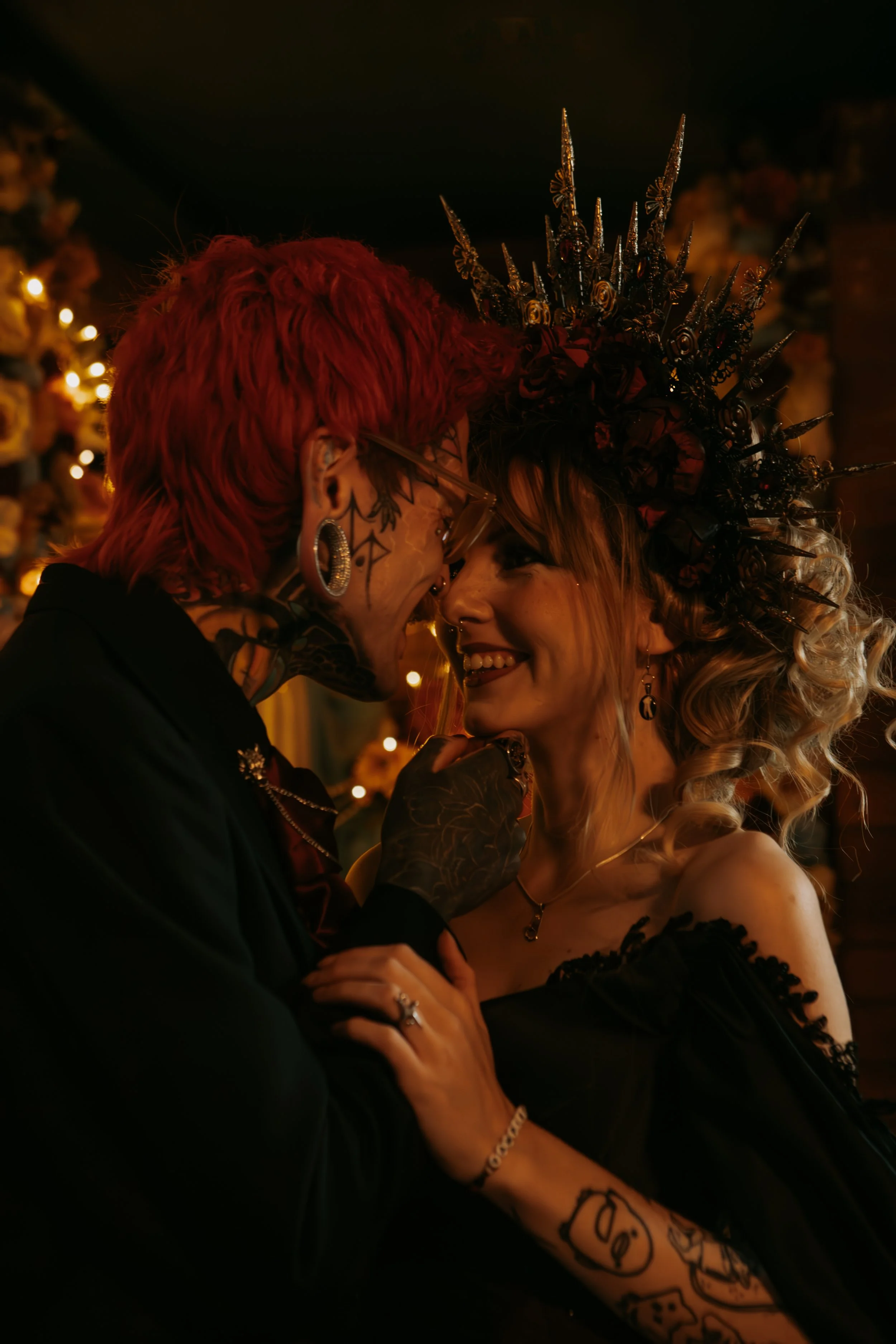 A couple, a man with pink hair and tattoos, and a woman in a black dress with a crown, share a close, intimate moment smiling at each other.