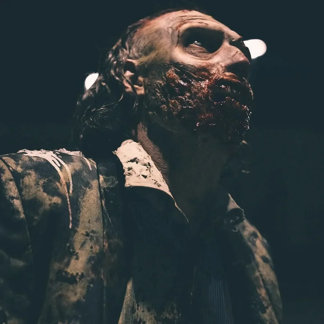 A close-up of a zombie with decomposed skin, dark eye sockets, and tattered clothing, in a dark setting.