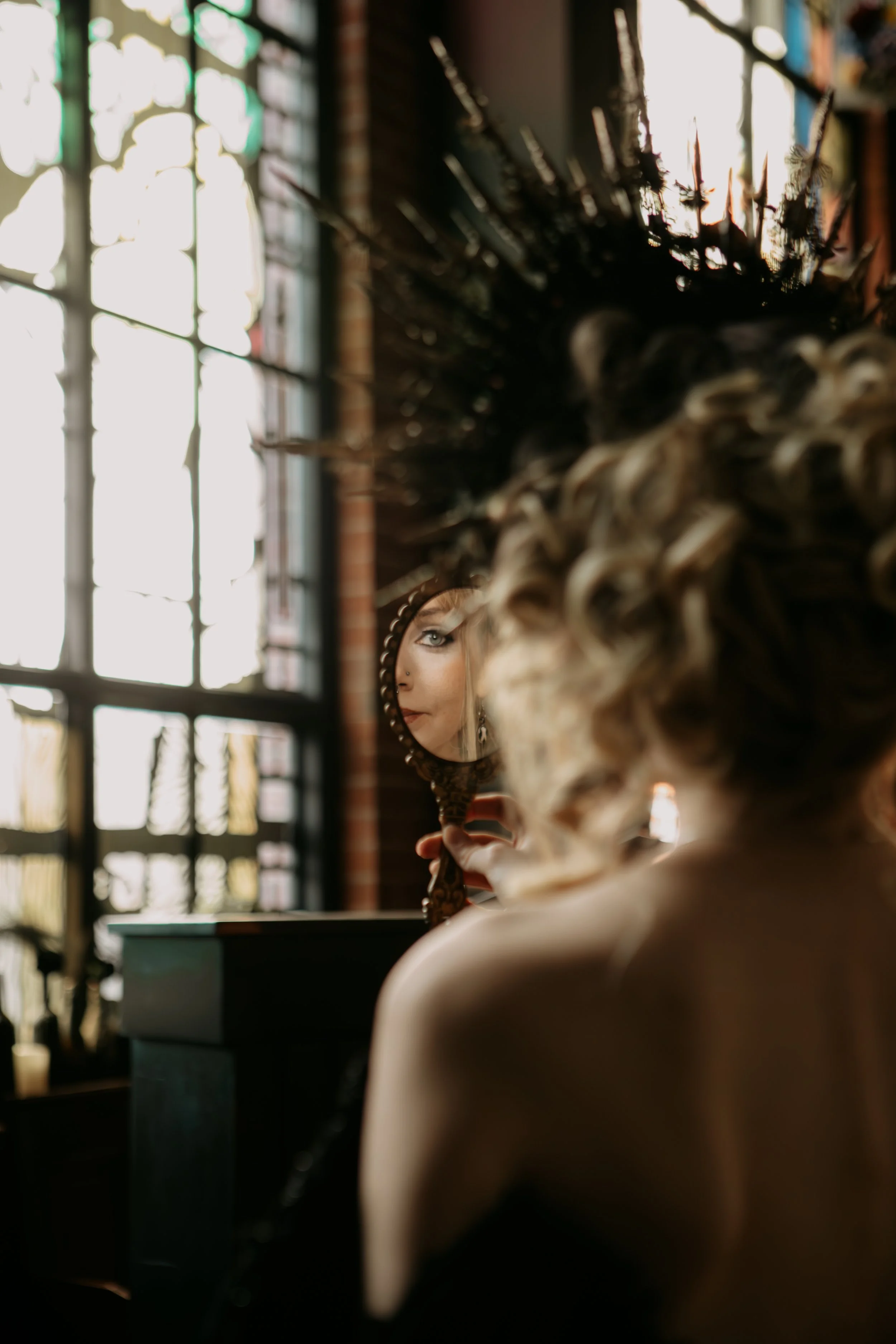 A woman with curly hair is holding a small mirror and looking at her reflection, situated in front of a large window with grid panes, inside a room with exposed brick walls.