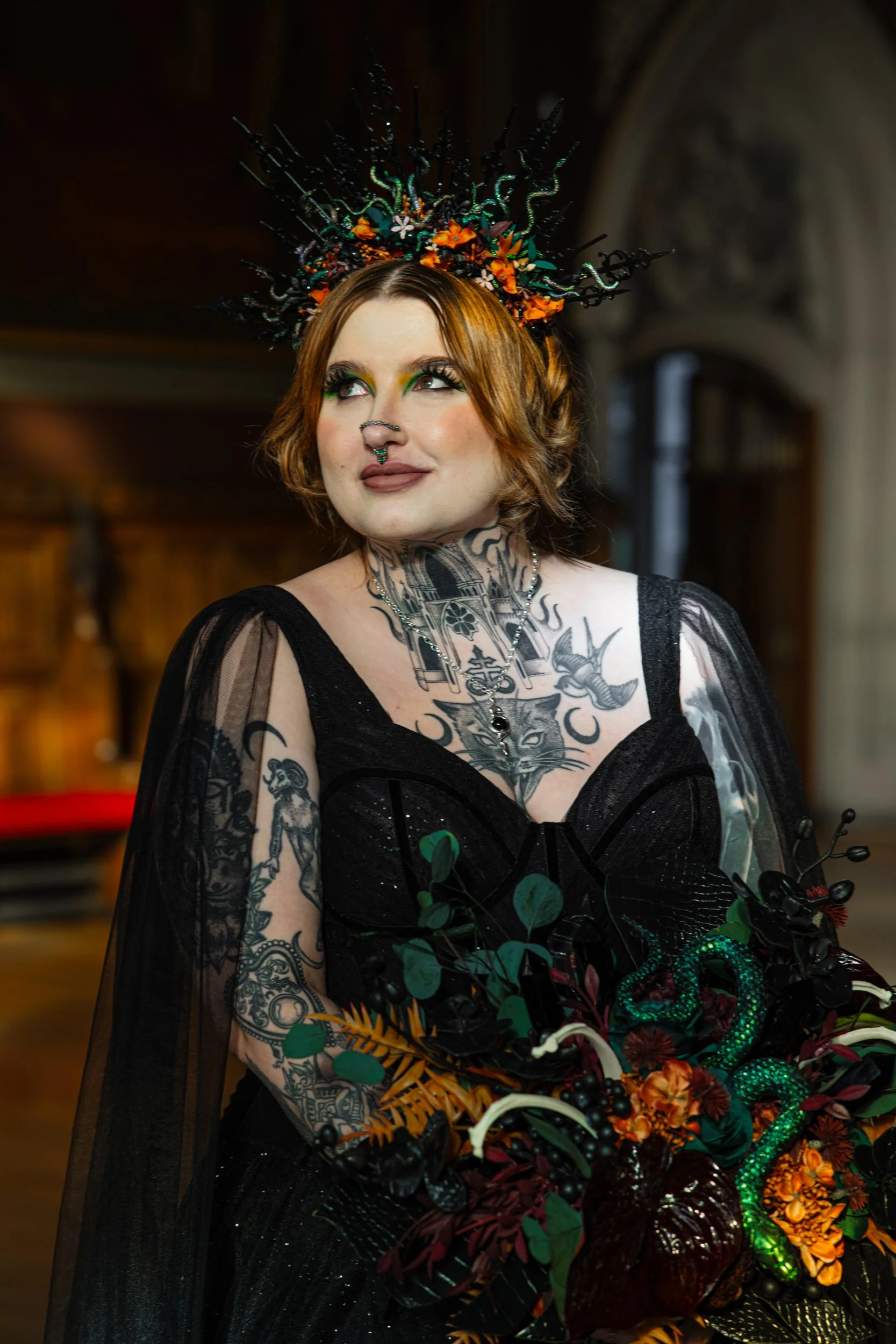 A woman with tattoos, red hair, and gothic makeup wearing a black dress and a decorative crown, holding a colorful bouquet with a snake sculpture.