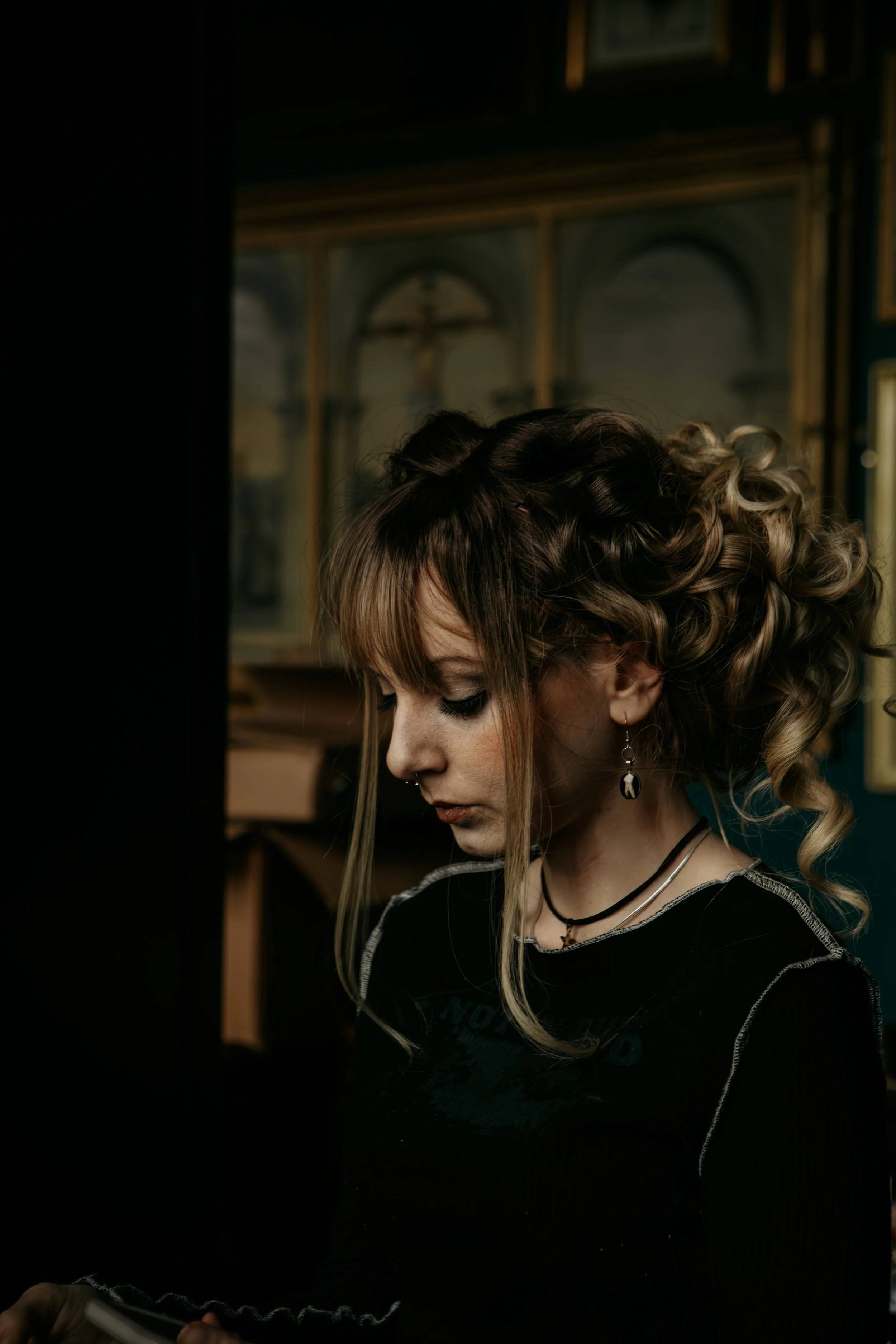 A woman with curled hair looking down, wearing earrings, necklaces, and a black top, in a dimly lit room with framed pictures and a crucifix in the background.
