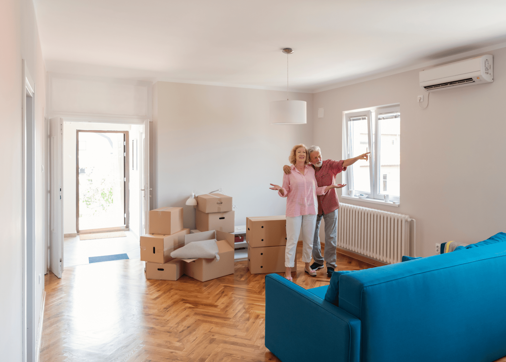 A happy senior couple standing inside a living room with moving boxes, smiling and gesturing happily.