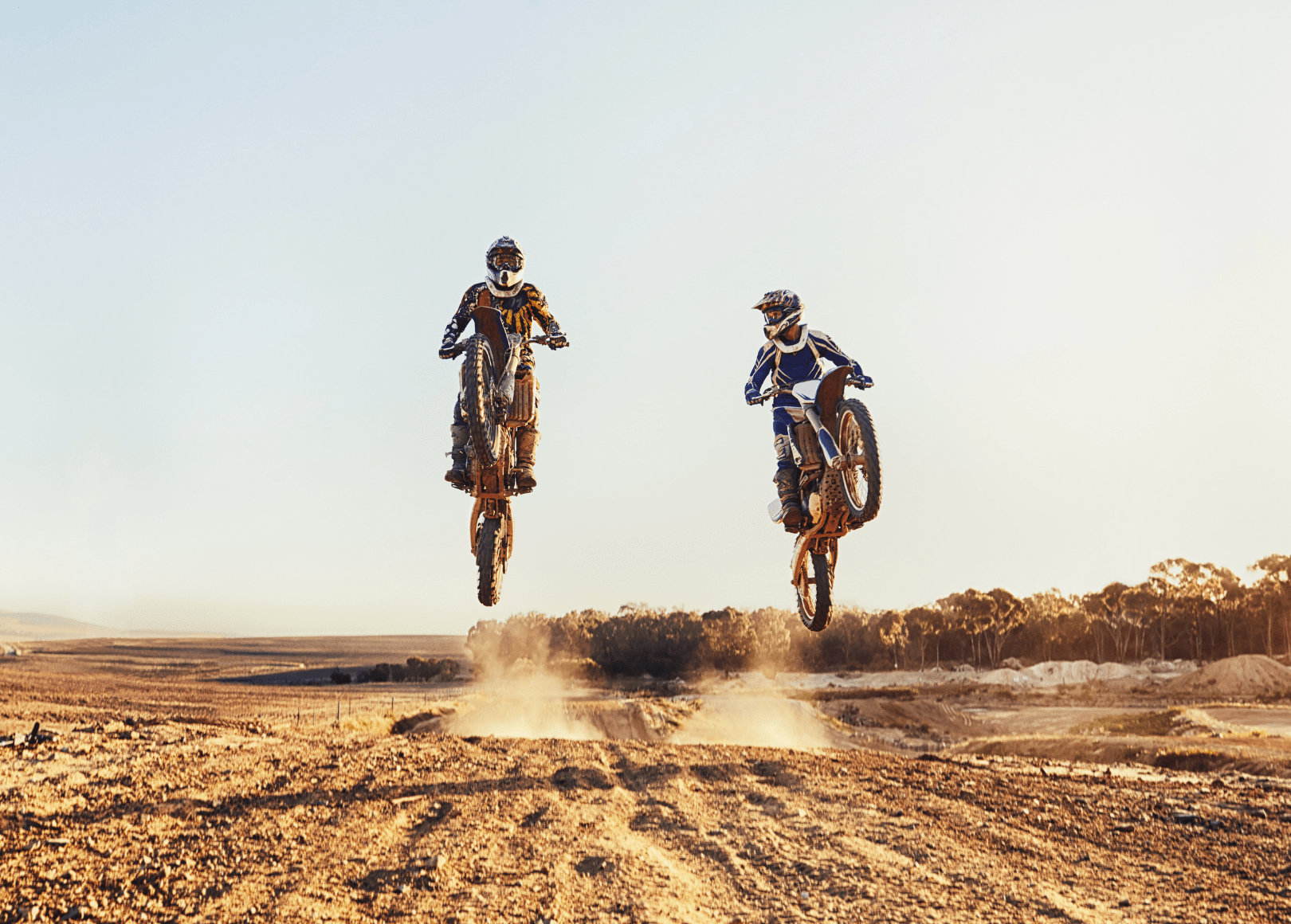 Two motocross riders performing wheelies in a desert landscape during sunset.